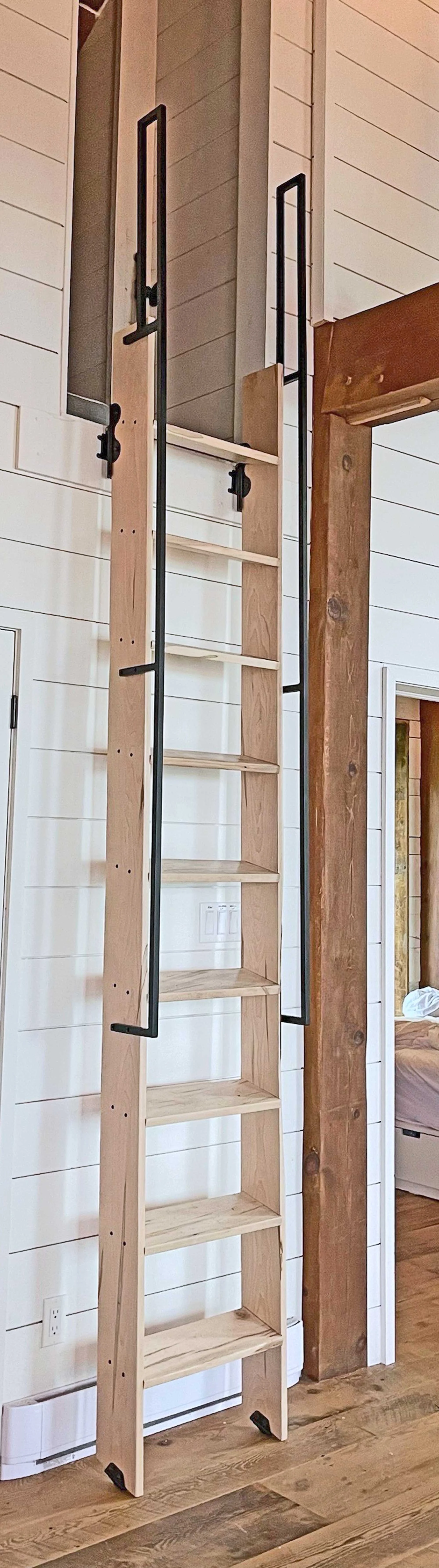 A tall wooden ladder with black metal rails leaning against a wall in a room with wooden floors and white shiplap walls.