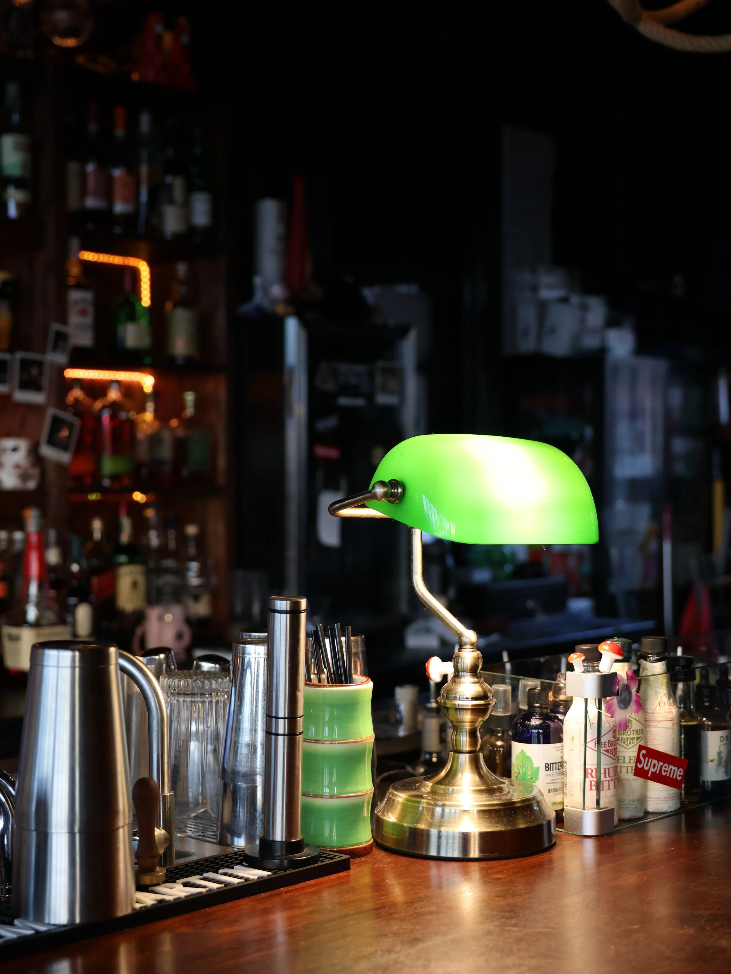 A bar scene with a green vintage desk lamp illuminating a wooden bar counter. On the counter are various bottles, a metal cocktail shaker, and a cup with black straws. In the background, shelves hold liquor bottles and small photographs, with warm st