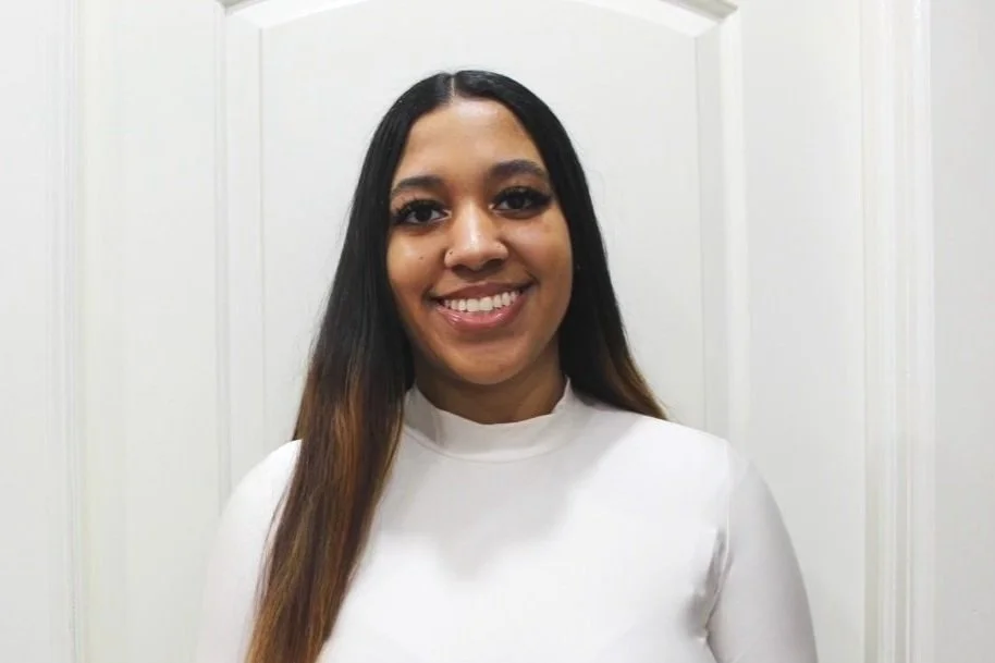 A woman with long dark hair and a white top, smiling in front of a white door.