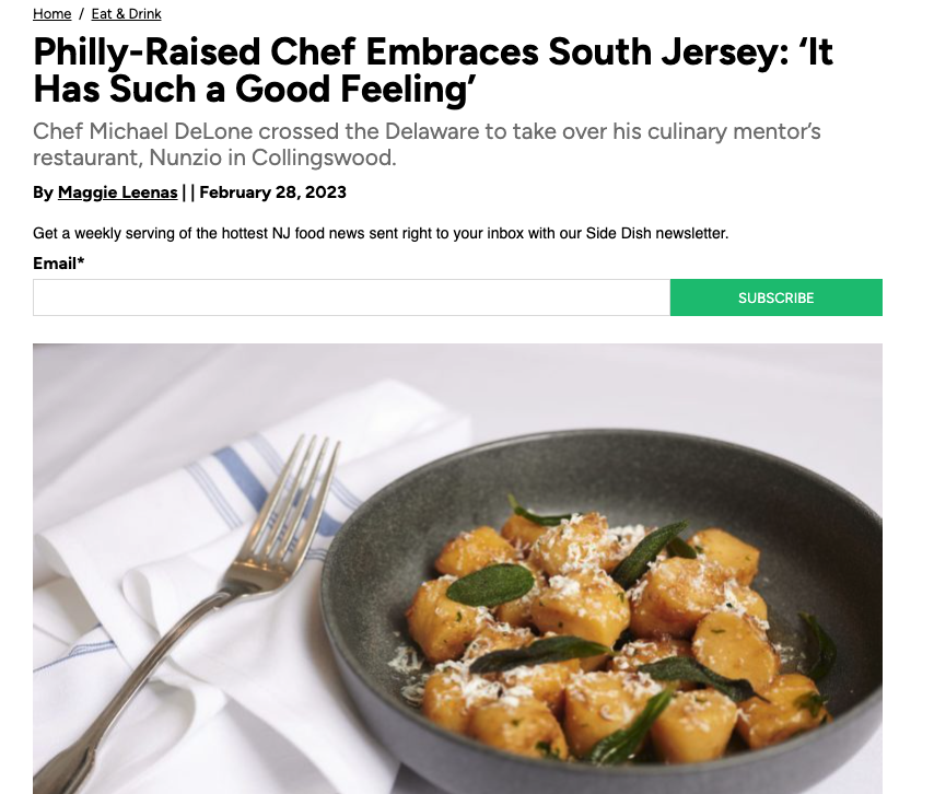 A black bowl filled with gnocchi topped with cheese and green leaves, placed beside a silver fork on a white napkin with blue stripes, on a white surface.