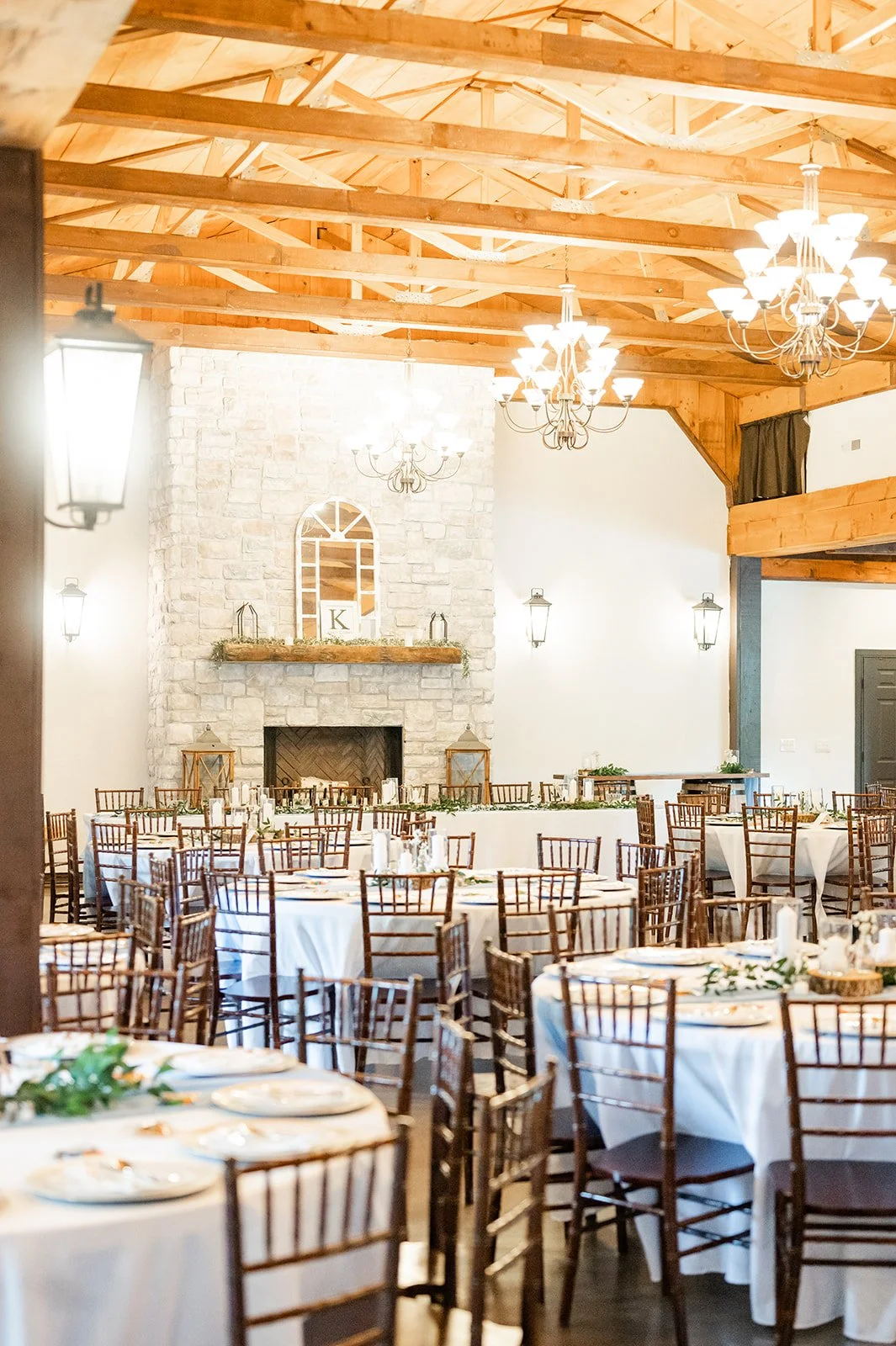 Indoor wedding reception with round tables covered in white tablecloths, wooden chairs, and a stone fireplace in a rustic barn with exposed wood beams and chandeliers.