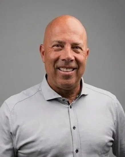 A smiling bald man wearing a light gray button-up shirt against a gray background.