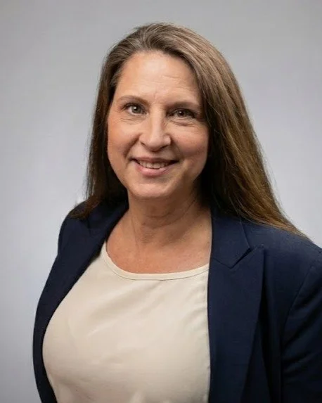 A woman with long brown hair smiling, wearing a dark blazer over a light-colored blouse against a plain gray background.