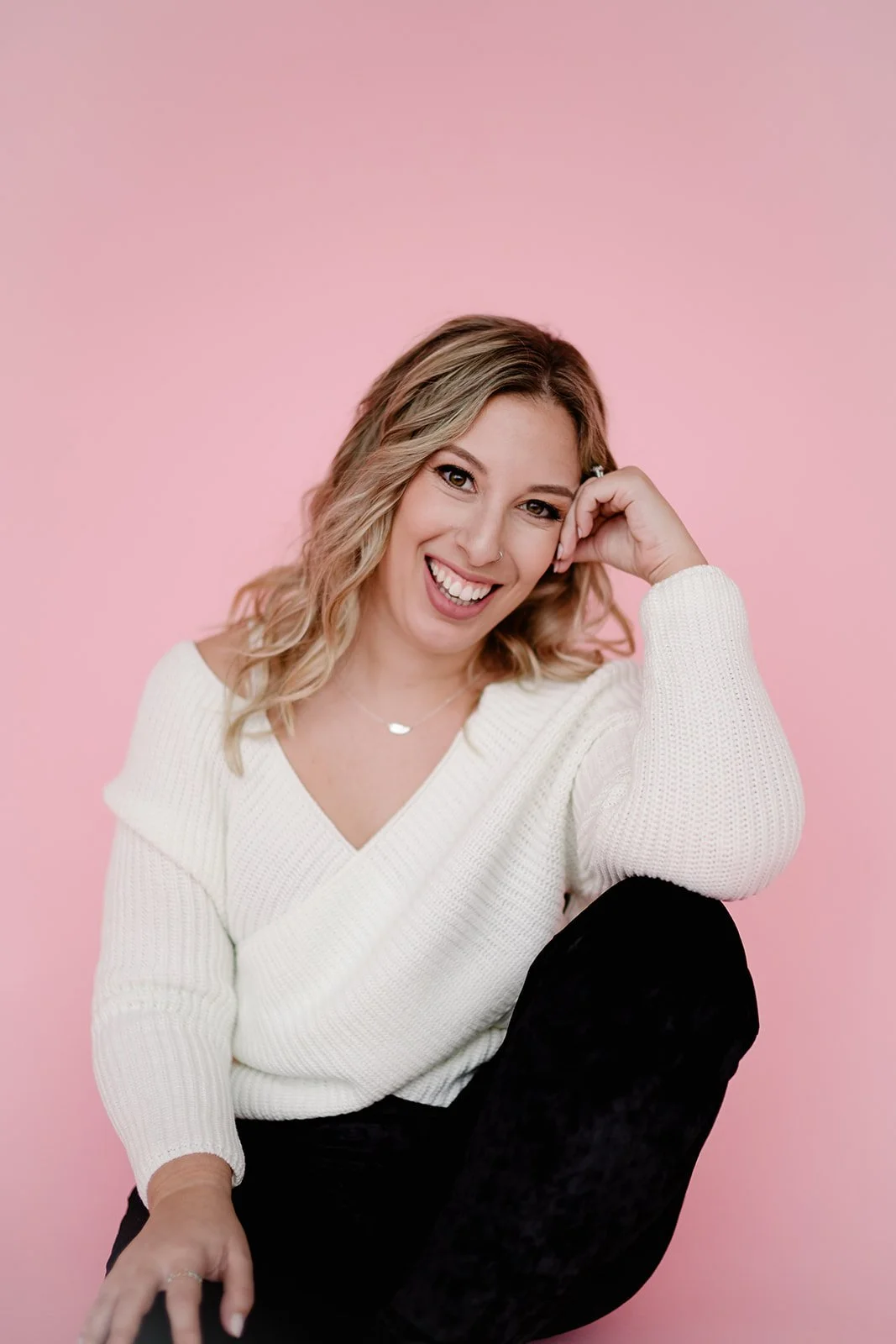 A young woman with wavy blonde hair, smiling, wearing a light blue dress with puffed sleeves, standing indoors against a white wall.