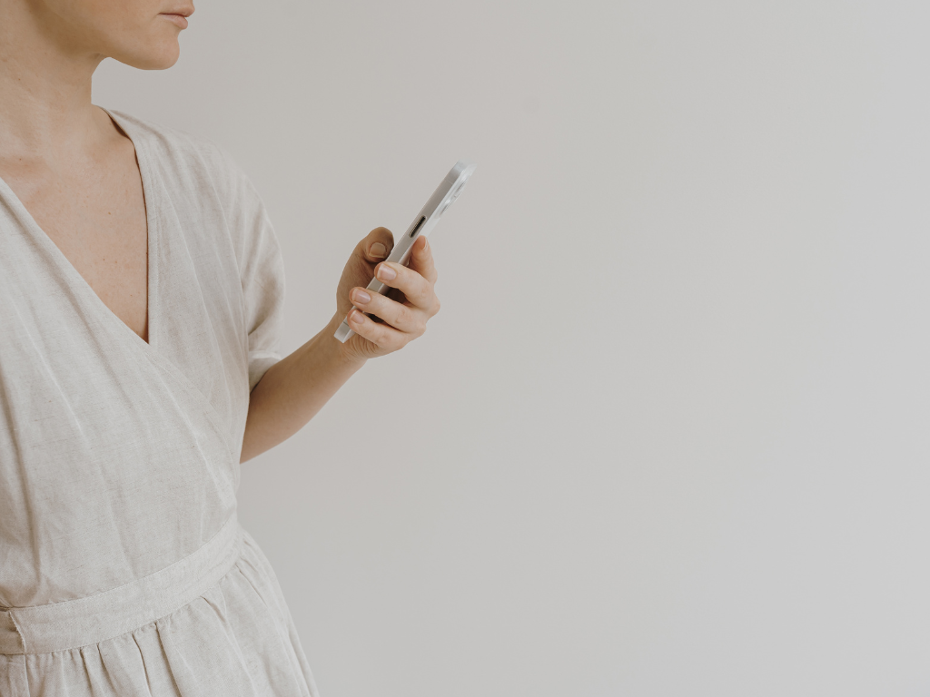 A woman wearing a beige dress holding a smartphone in her right hand, standing against a plain white wall.