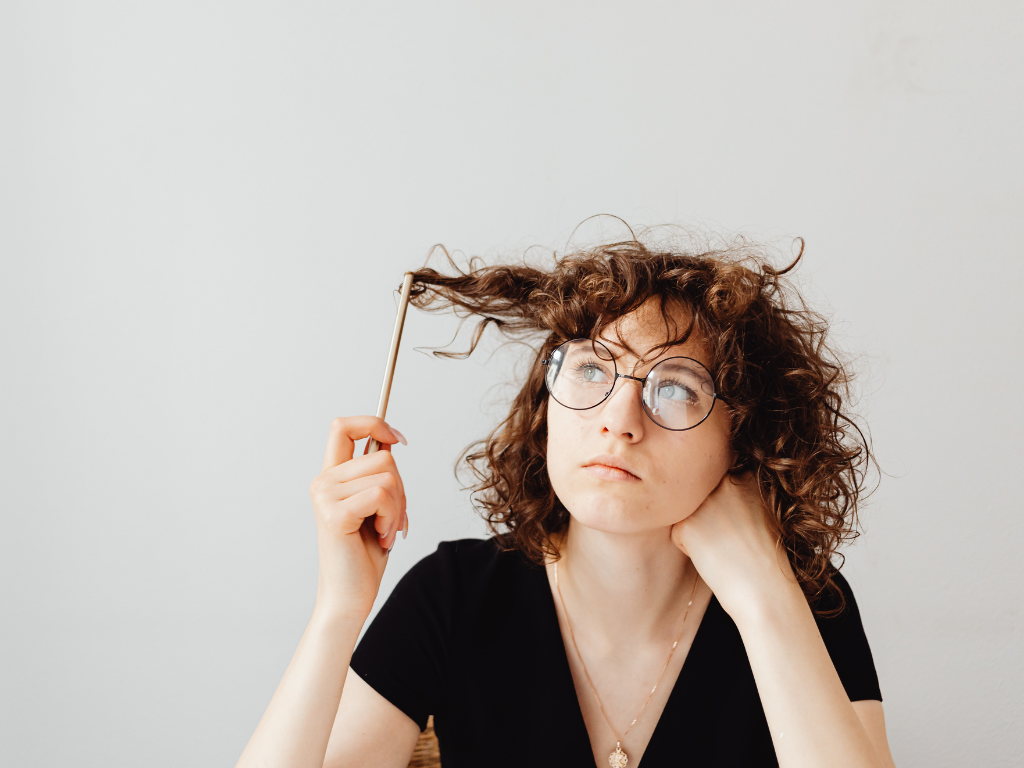 Young woman with curly hair and glasses holding a strand of her hair, looking thoughtul.