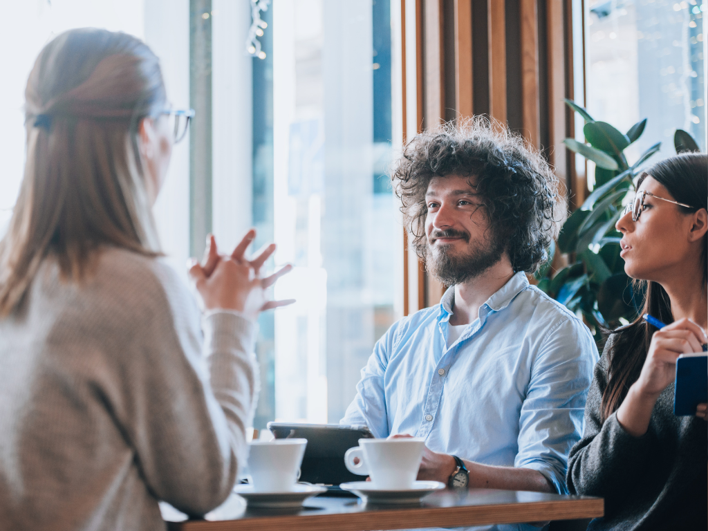 Three people having a discussion at a coffee shop, two women and a man with curly hair, with coffee cups on the table.
