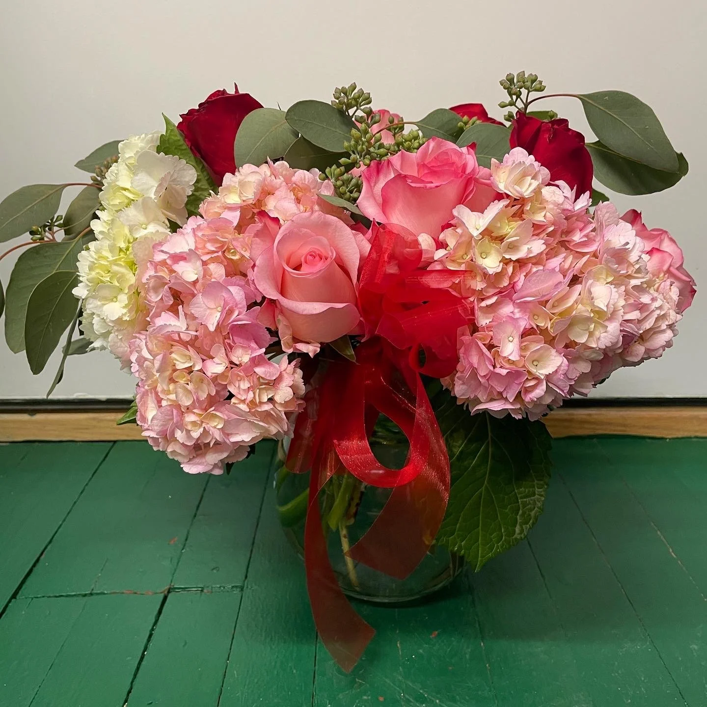 Floral arrangement with pink roses, hydrangeas, red roses, white flowers, and green leaves in a glass vase on a green floor.