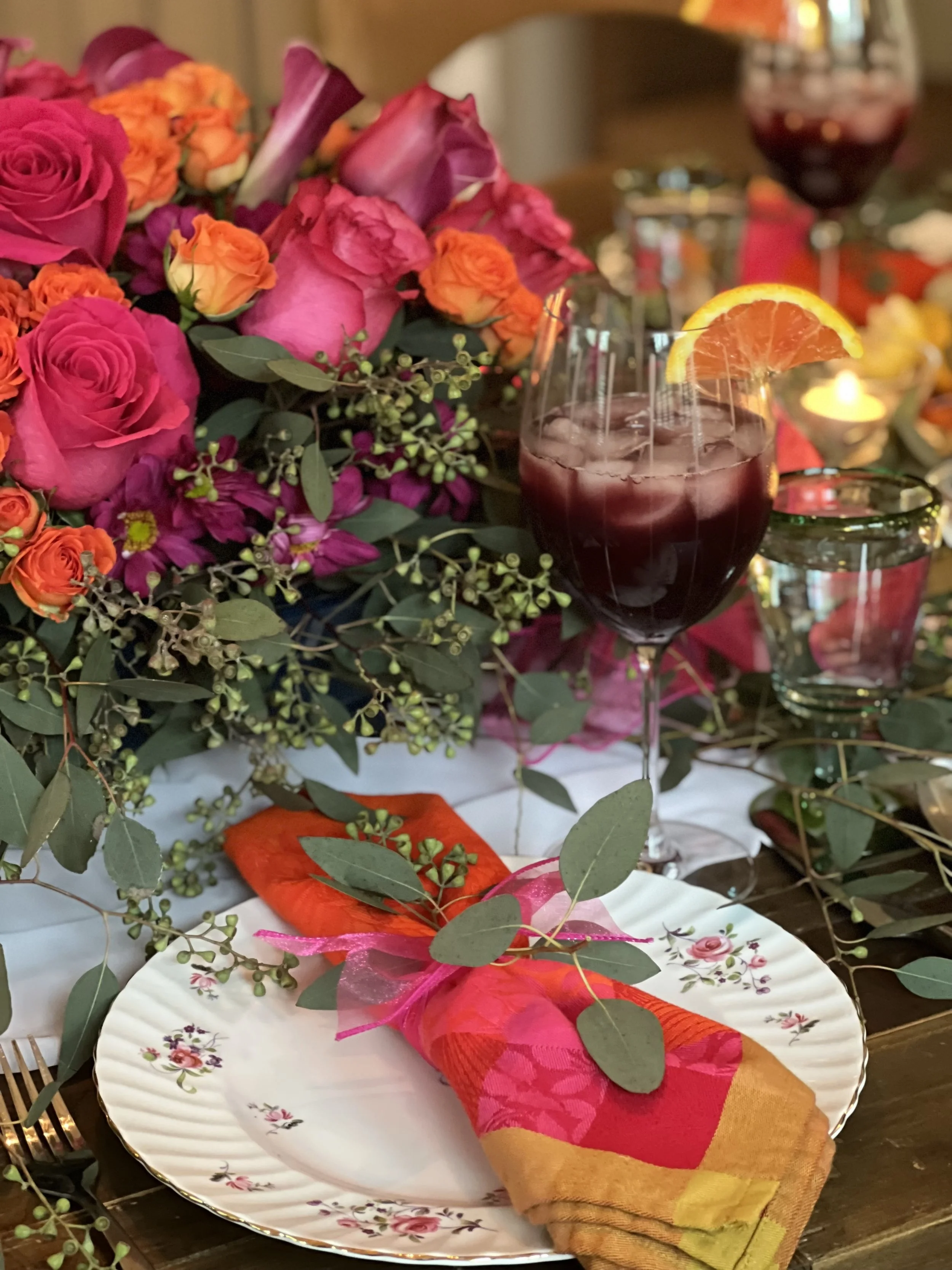 Elegant table setting with floral arrangement featuring pink and orange roses, greenery, and a glass of sangria garnished with an orange slice.