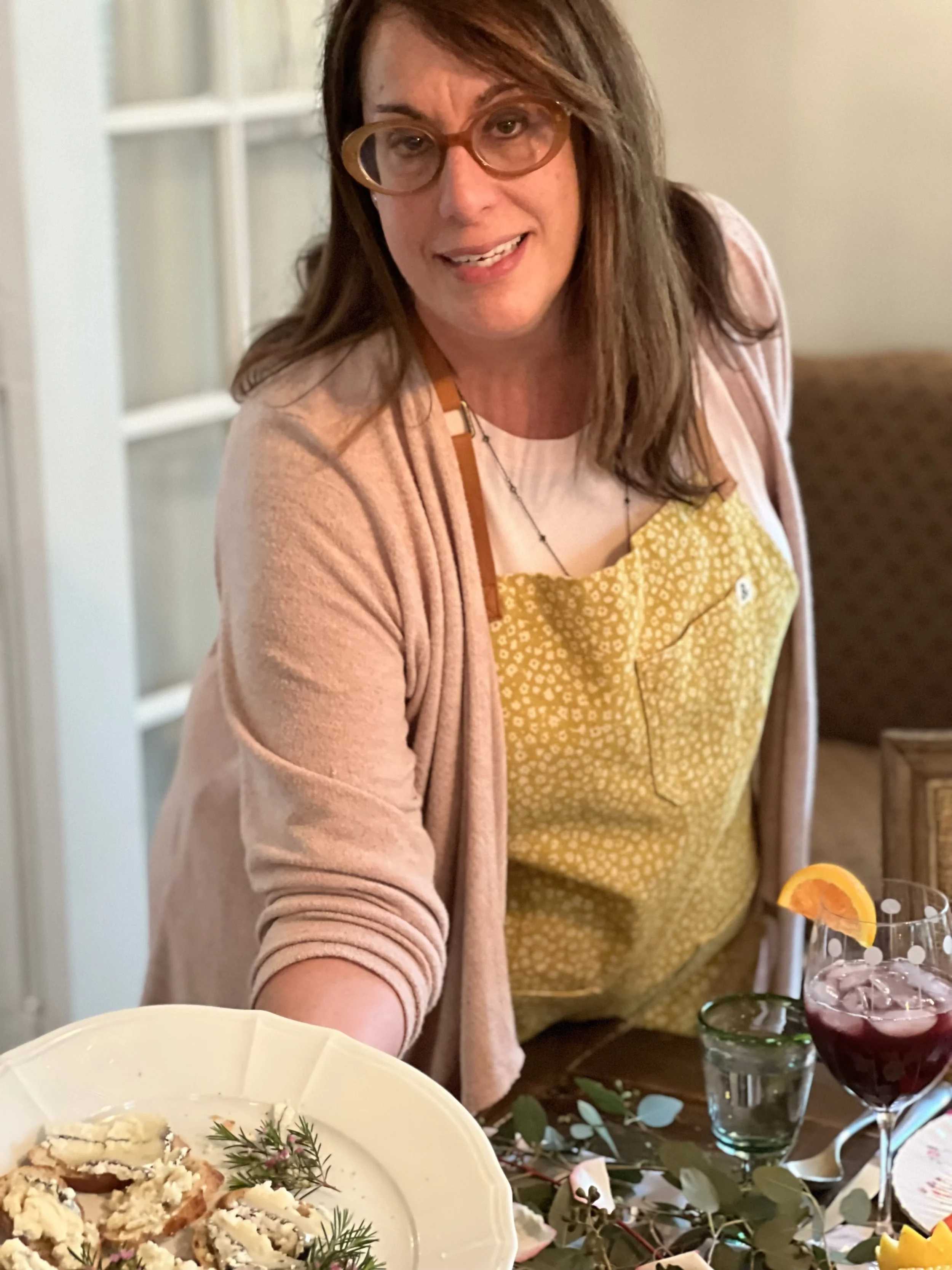 Woman wearing glasses and a yellow apron serving a plate of appetizers, with a drink on the table.