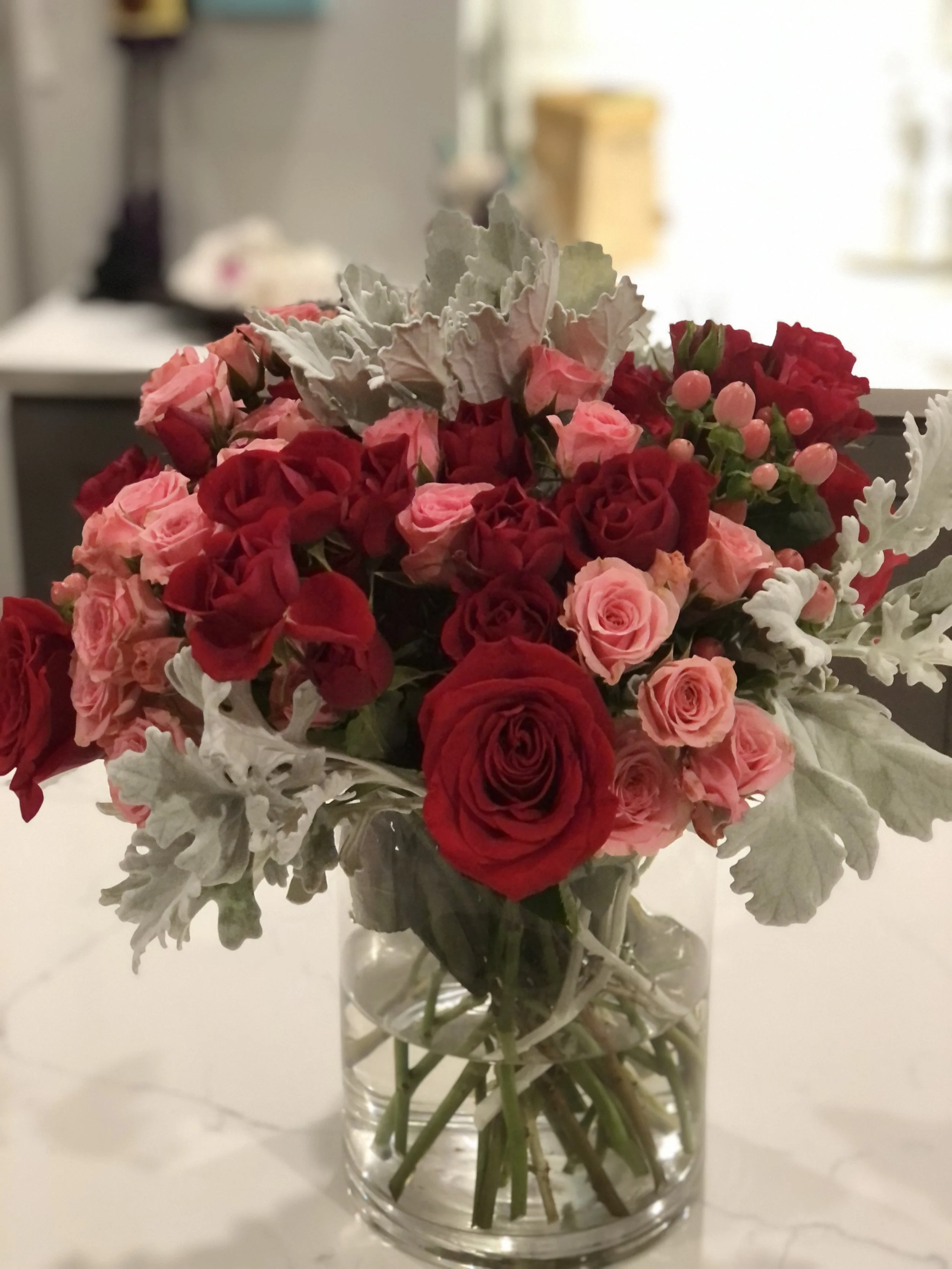 Bouquet of red and pink roses with dusty miller leaves in a clear glass vase on a white surface.