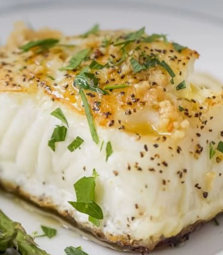 Baked white fish fillet topped with garlic and herbs, served with asparagus on a white plate.