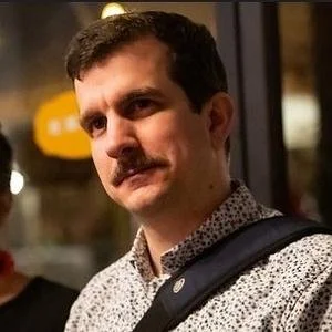 A man with dark hair and a mustache in a patterned shirt, carrying a backpack, standing indoors with a serious expression.