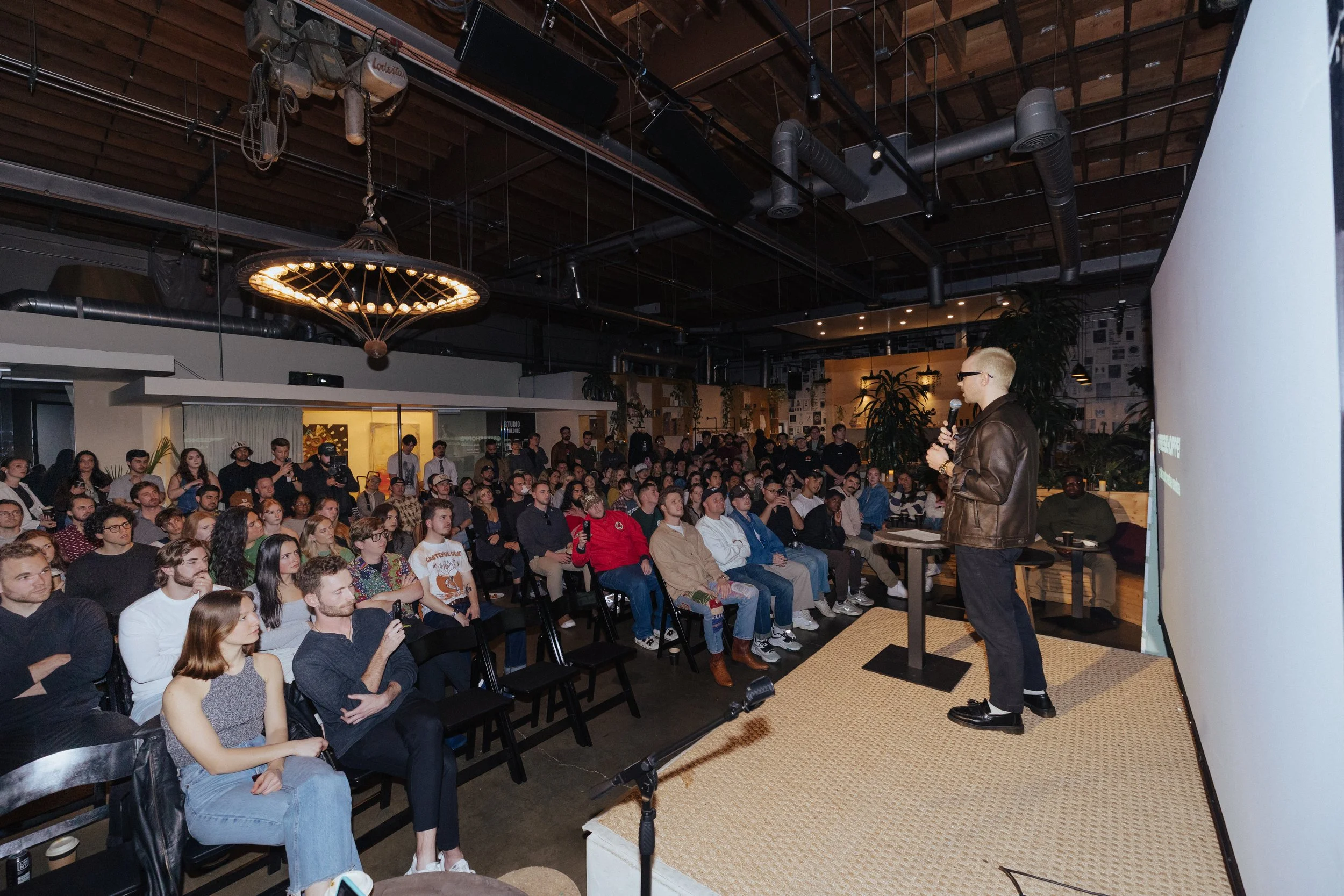 A man in a leather jacket giving a presentation to a large audience in a dimly lit conference room with plants and artwork on the walls.