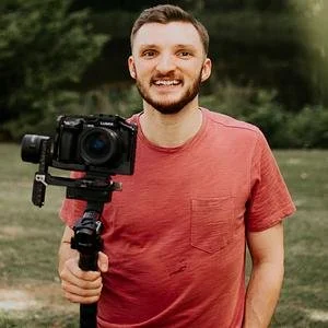 A smiling young man holding a camera mounted on a stabilizer outdoors in a park.