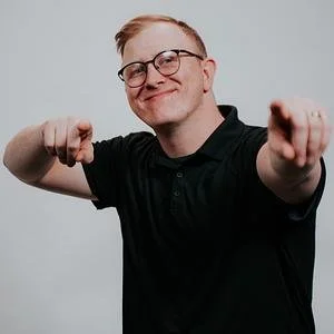 A young man wearing glasses, smiling confidently, pointing towards the camera with both hands, against a plain background.