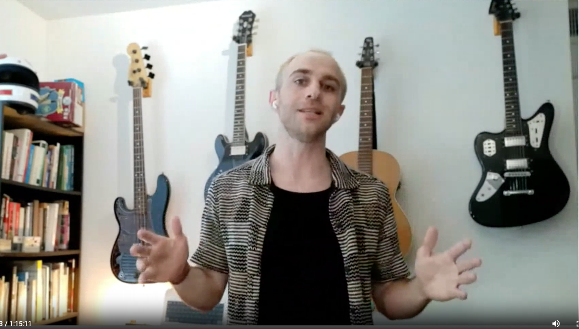 Man speaking in front of a wall with four electric guitars hanging and a bookshelf filled with books to the left.