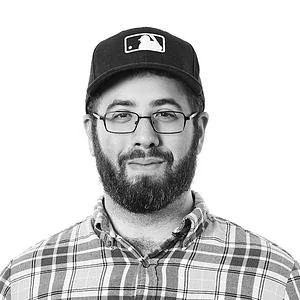 A man with glasses, a beard, and a mustache wearing a baseball cap and a plaid shirt, smiling slightly.