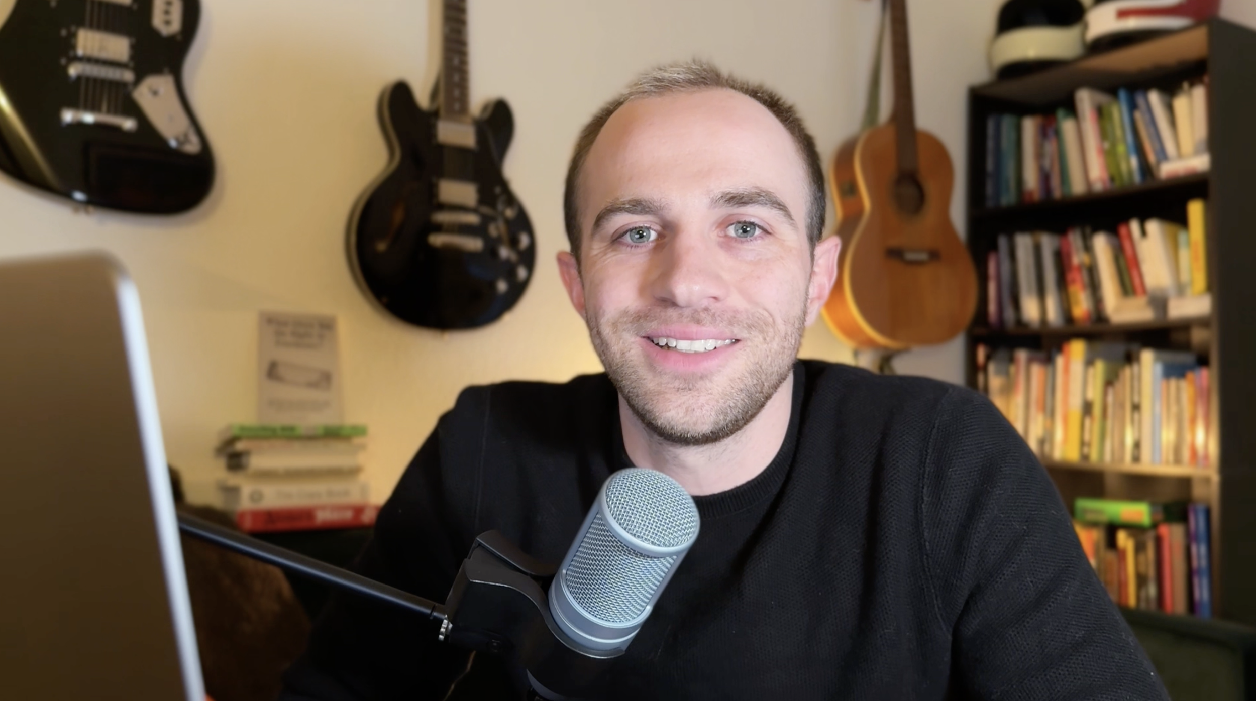 A man with short brown hair and a beard smiling at the camera, sitting in front of a microphone. Behind him are guitars hanging on the wall and a bookshelf filled with books.