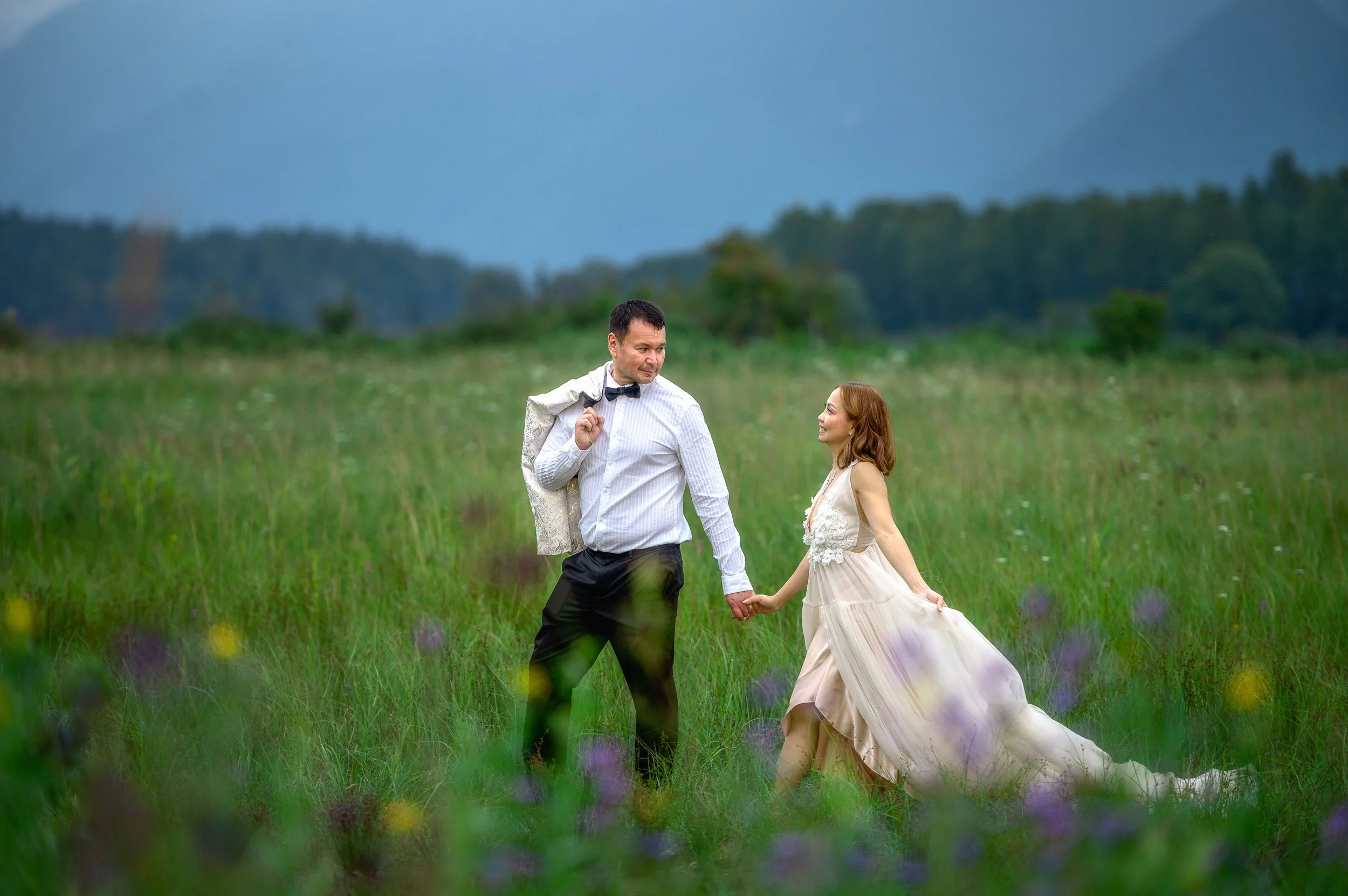 A man and woman holding hands in a grassy field, with mountains in the background, the man wearing a suit with a jacket draped over his shoulder and the woman in a flowing dress.