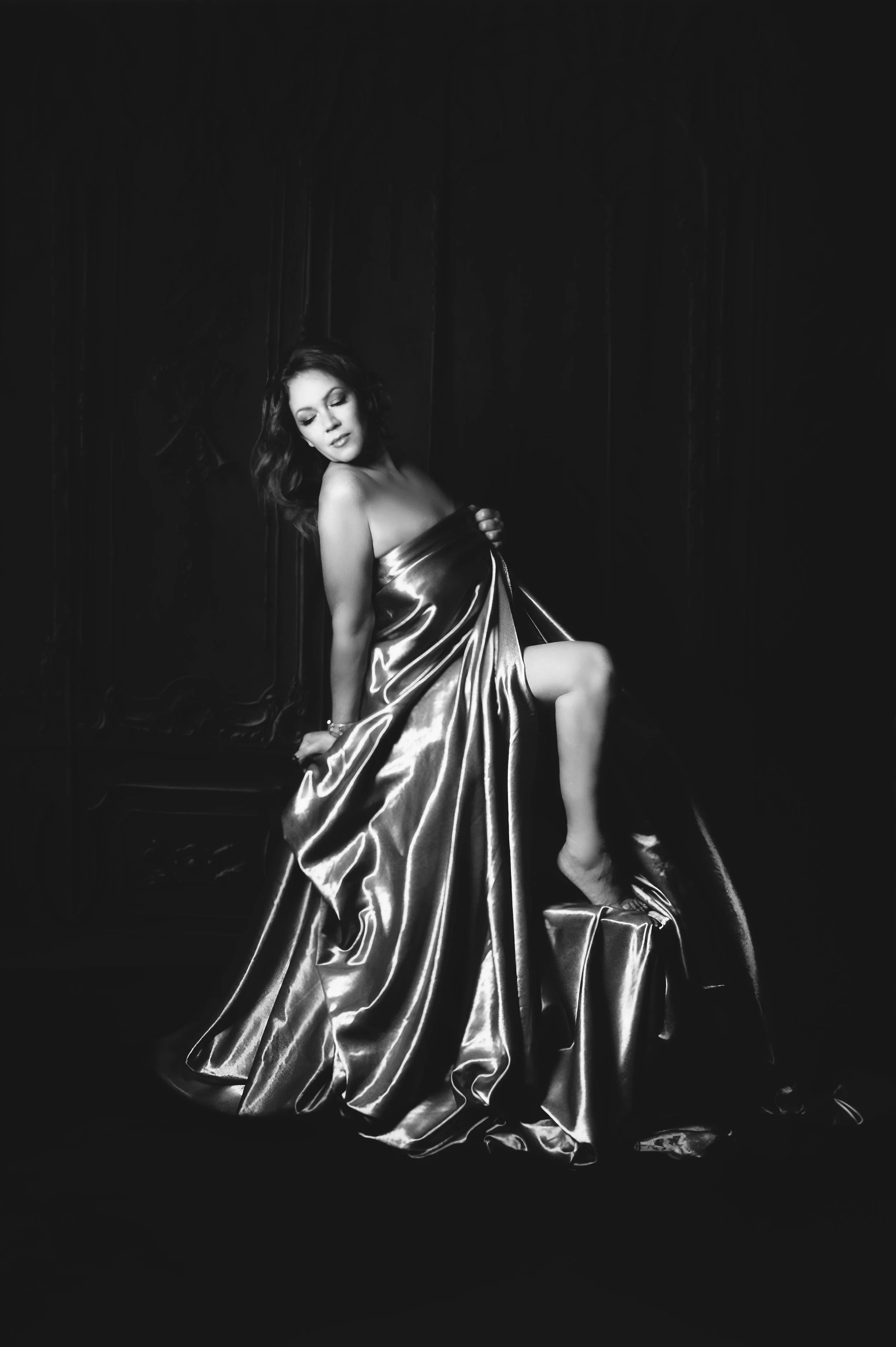 A black and white photograph of a woman in a metallic dress, sitting on a chair, with one leg raised and eyes closed, in a dark studio setting.