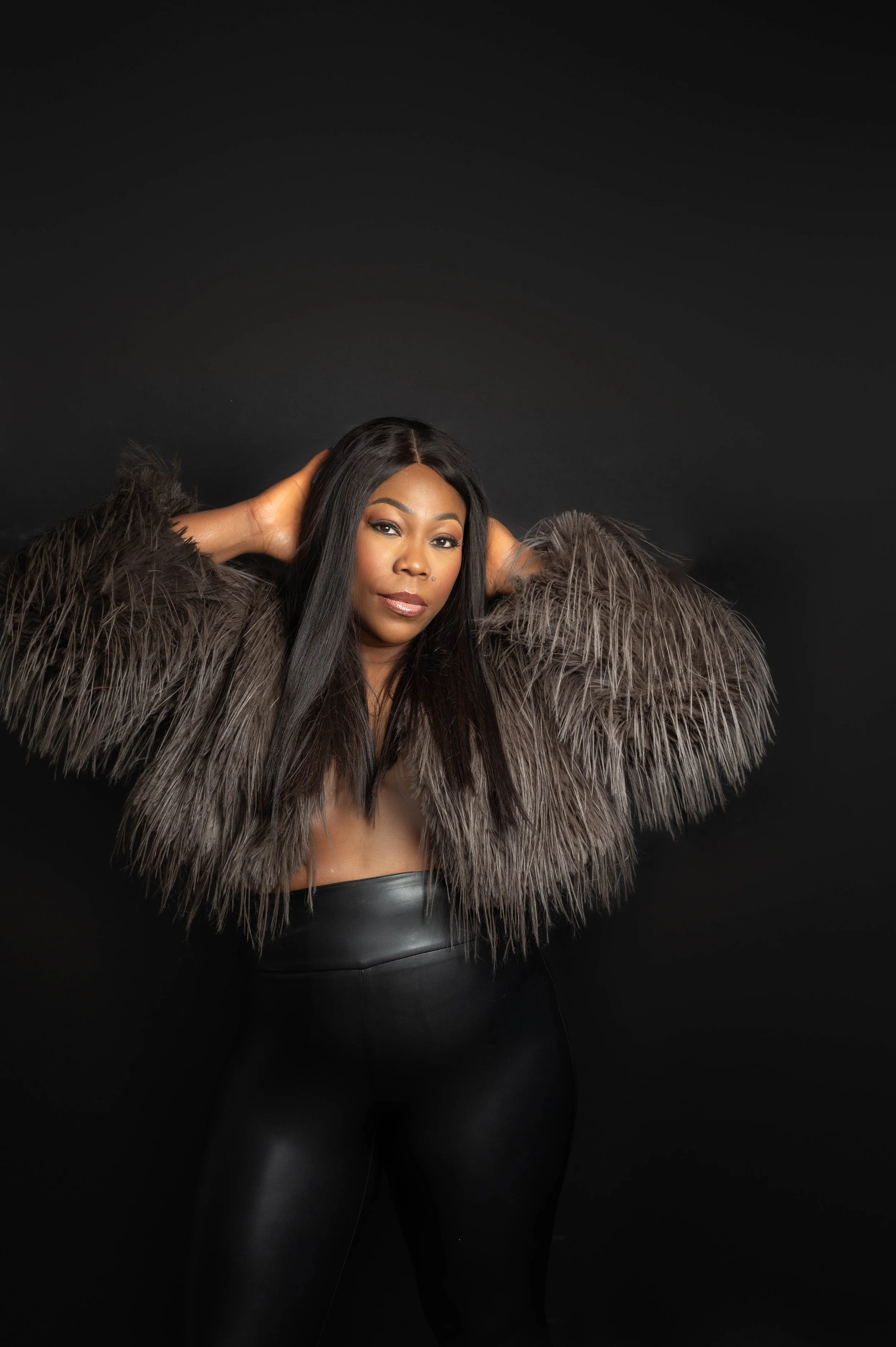 A woman wearing a feathered jacket and black leather pants, striking a pose against a black background.
