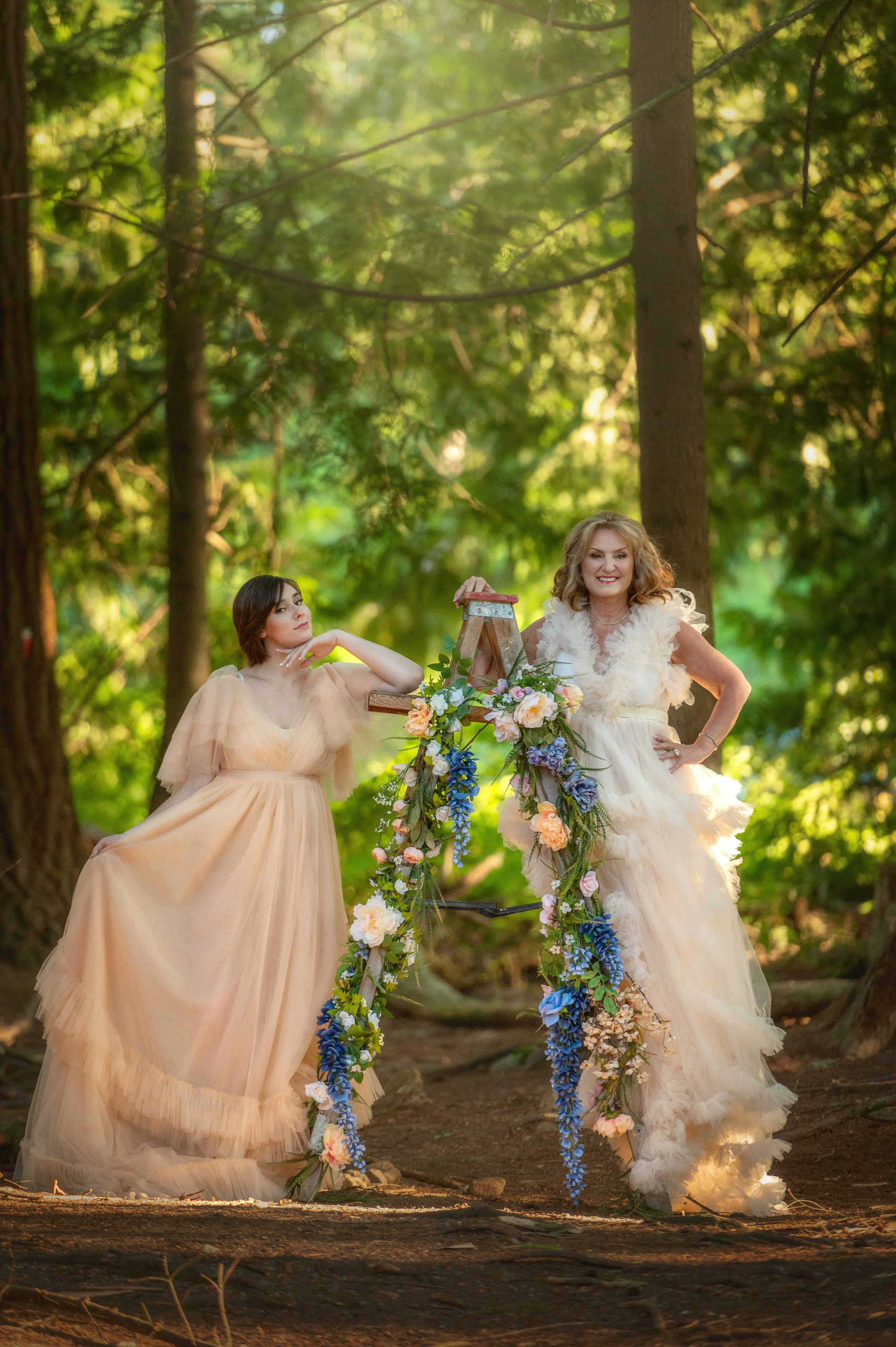 Two women in flowy dresses posing in a forest. One woman is leaning on a floral bicycle, and both are smiling.