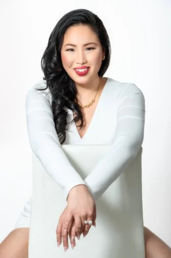 A woman with long black hair, wearing a white top and gold necklace, sitting on a white chair and smiling at the camera.