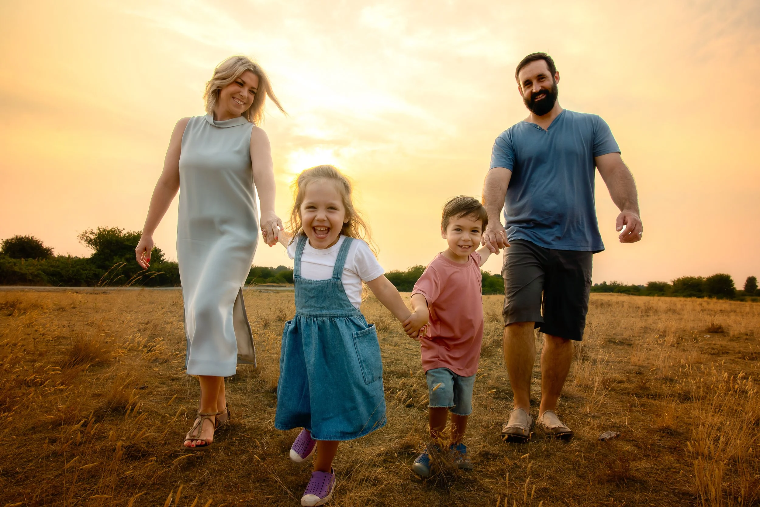 A family of four walking hand-in-hand on a grassy field at sunset, smiling and enjoying the outdoors.