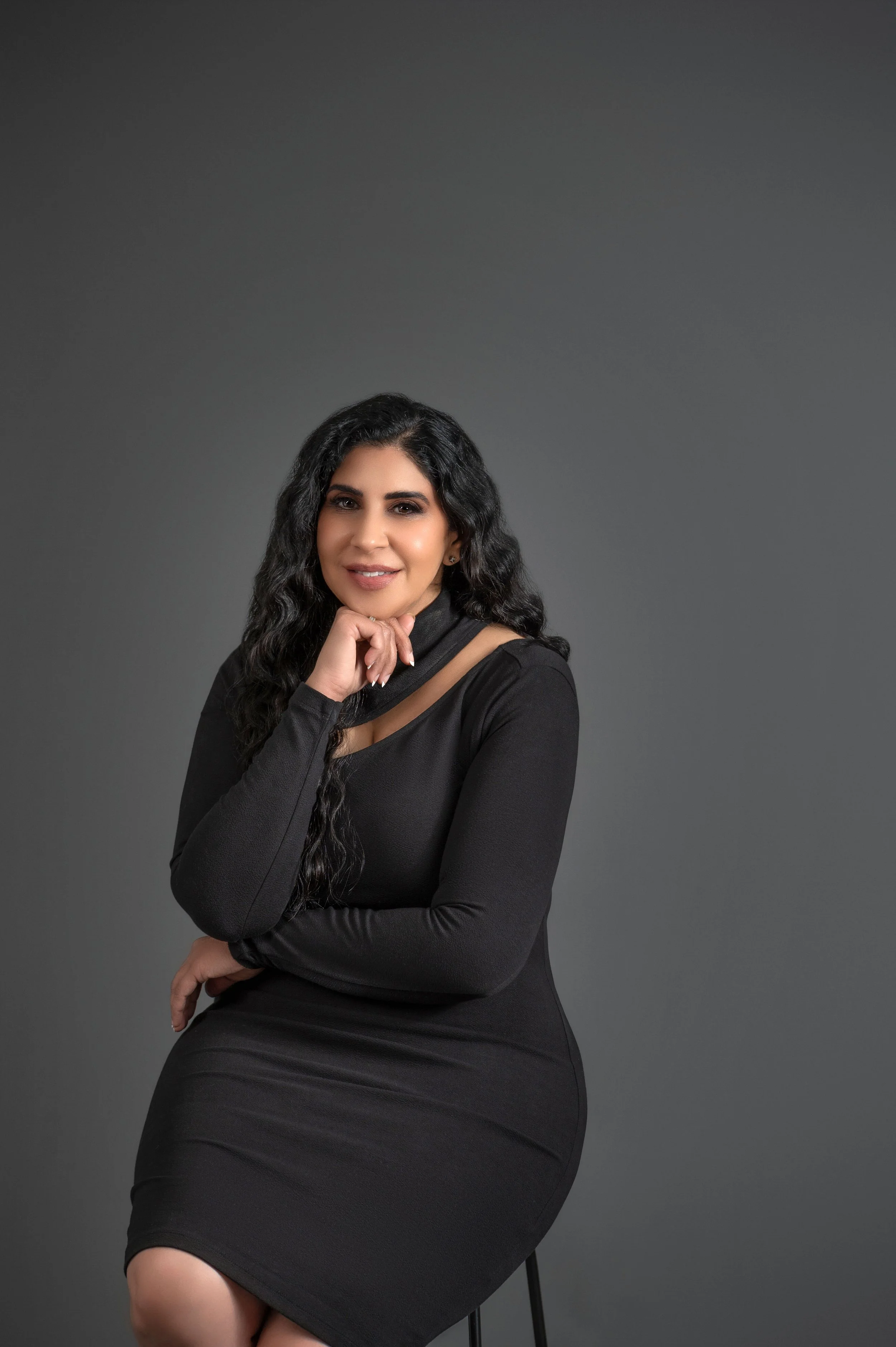 A woman with long curly black hair wearing a fitted black dress, sitting on a stool in front of a grey background, smiling and resting her chin on her hand.