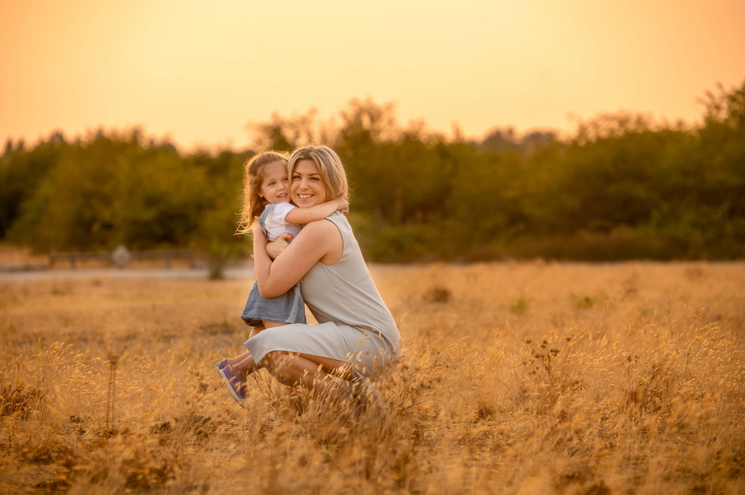 A woman and a young girl hugging in a field during sunset, both smiling happily.