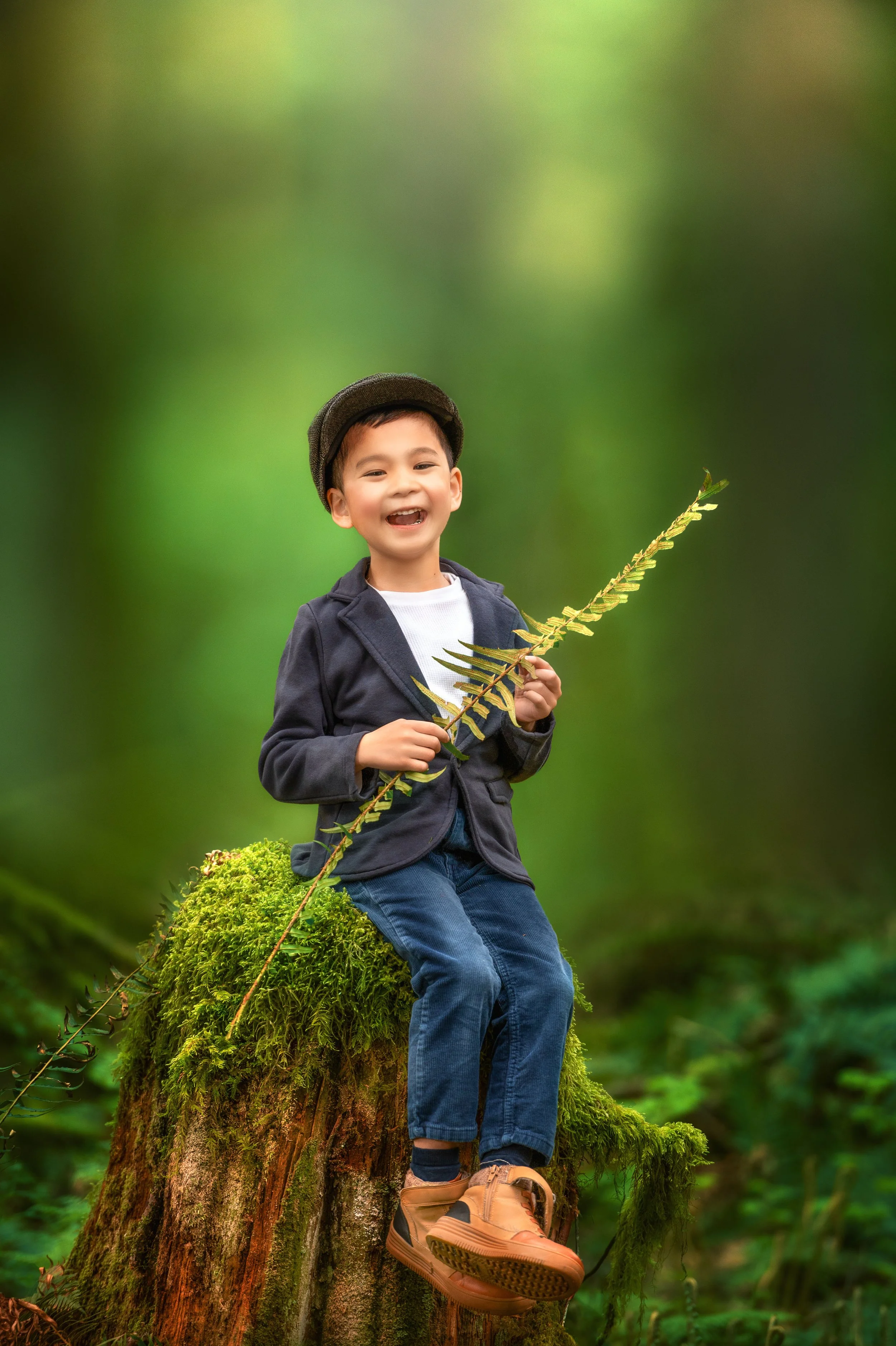 A young boy sitting on a moss-covered tree stump in a forest, smiling and holding a fern leaf.