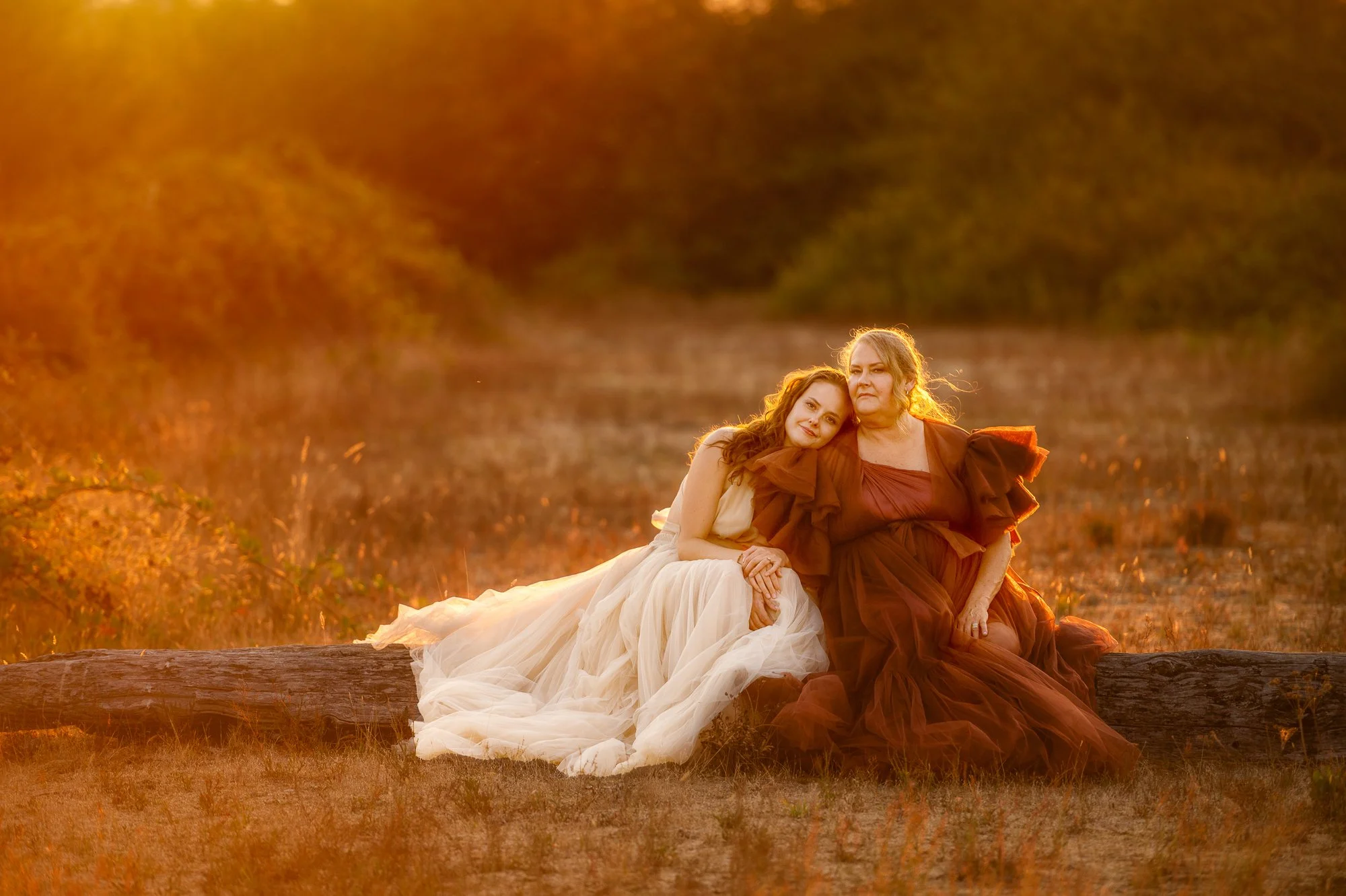 A young woman and an older woman sitting on a log outdoors during sunset, wearing flowing dresses, with the young woman leaning her head on the older woman's shoulder.