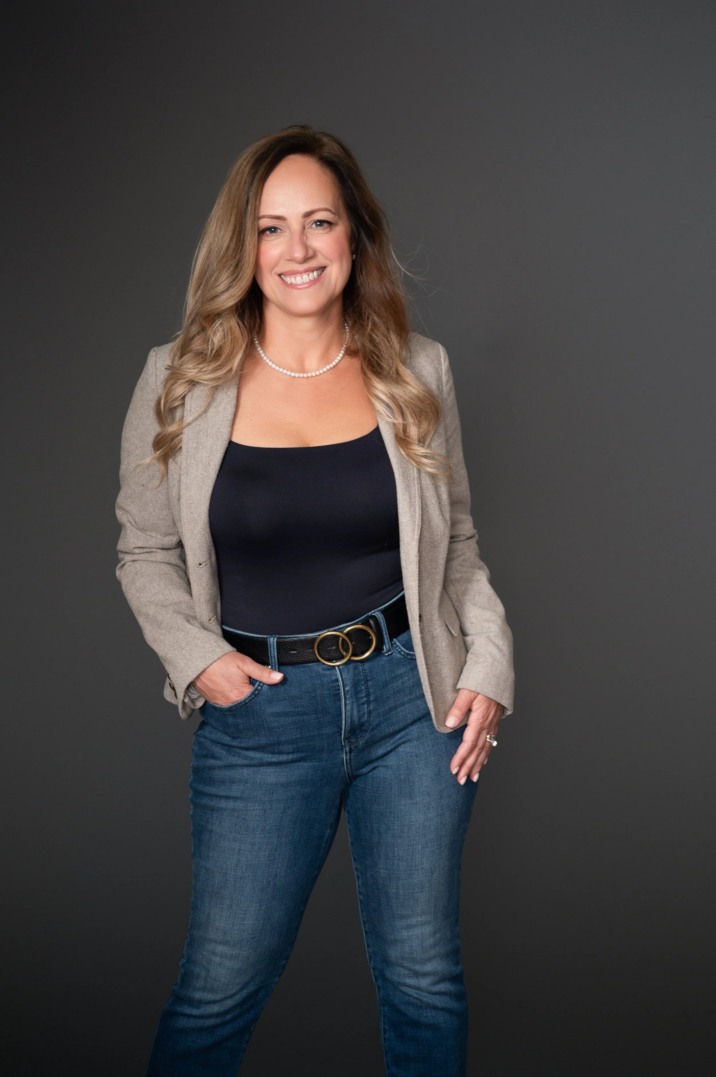 Woman with long wavy hair, wearing a beige blazer, black top, blue jeans, and a pearl necklace, standing against a dark gray background.