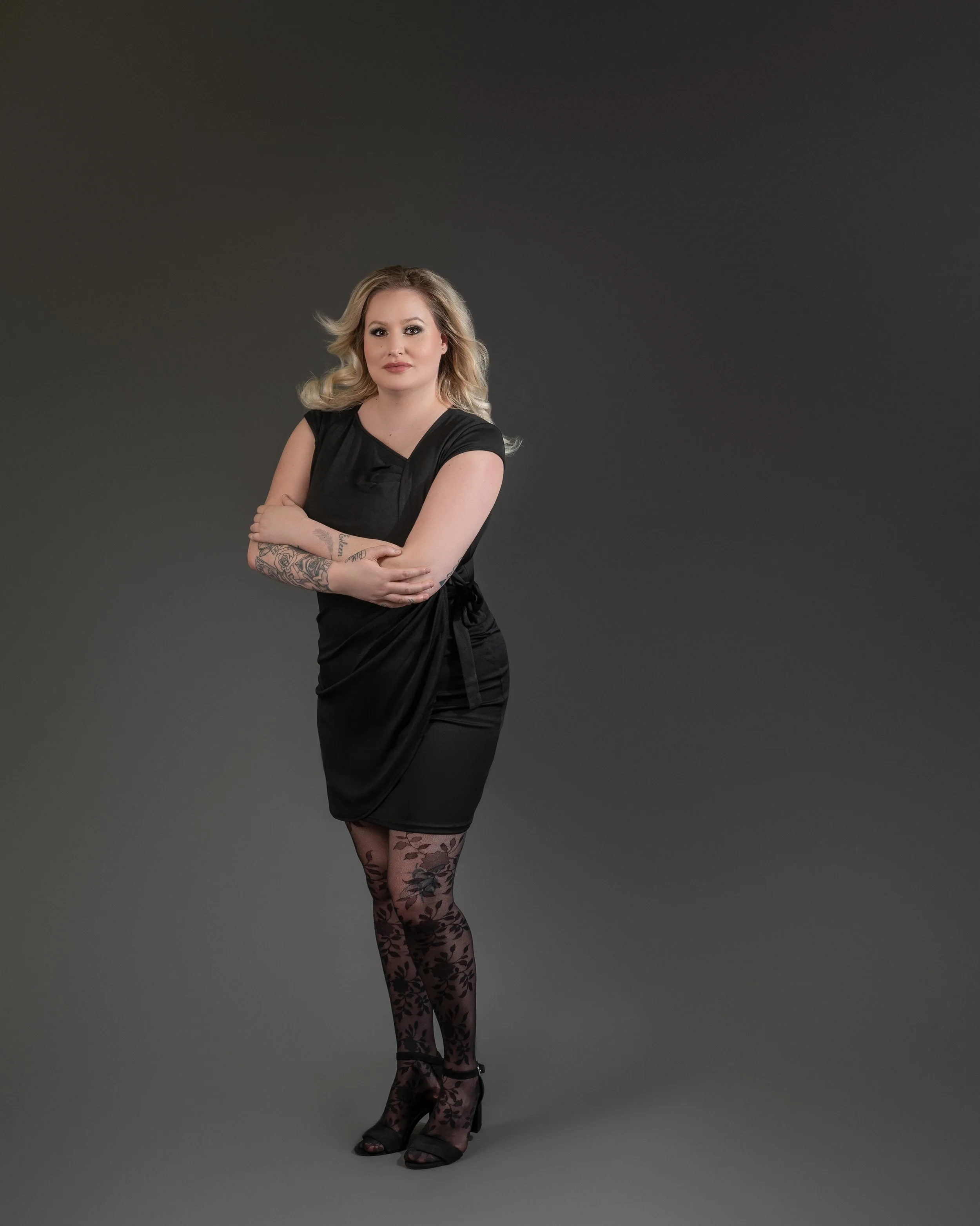 A woman with blonde curly hair wearing a black dress with lace tights and black heels.