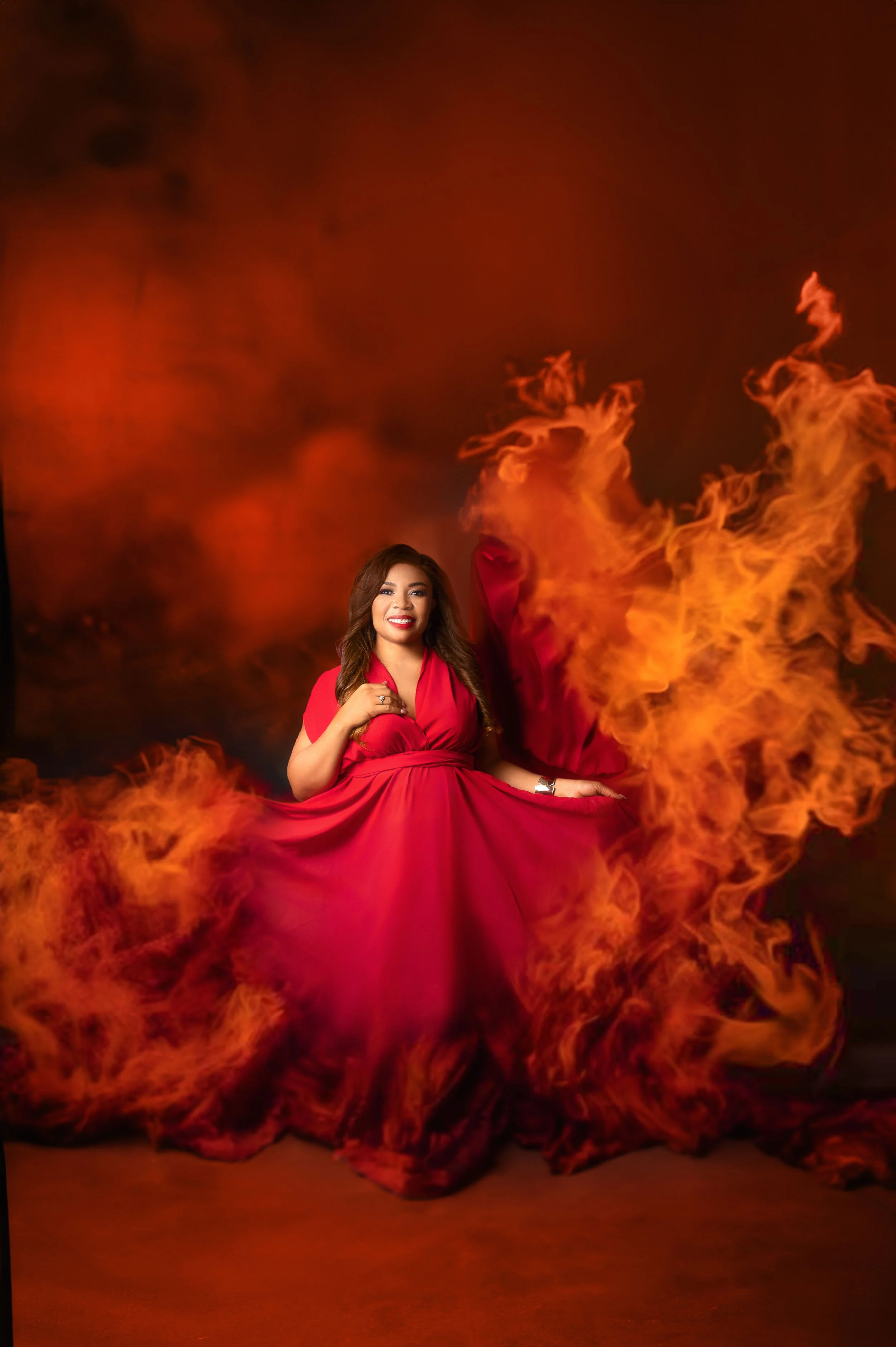 A smiling woman in a red dress sitting among flames with a dark background.