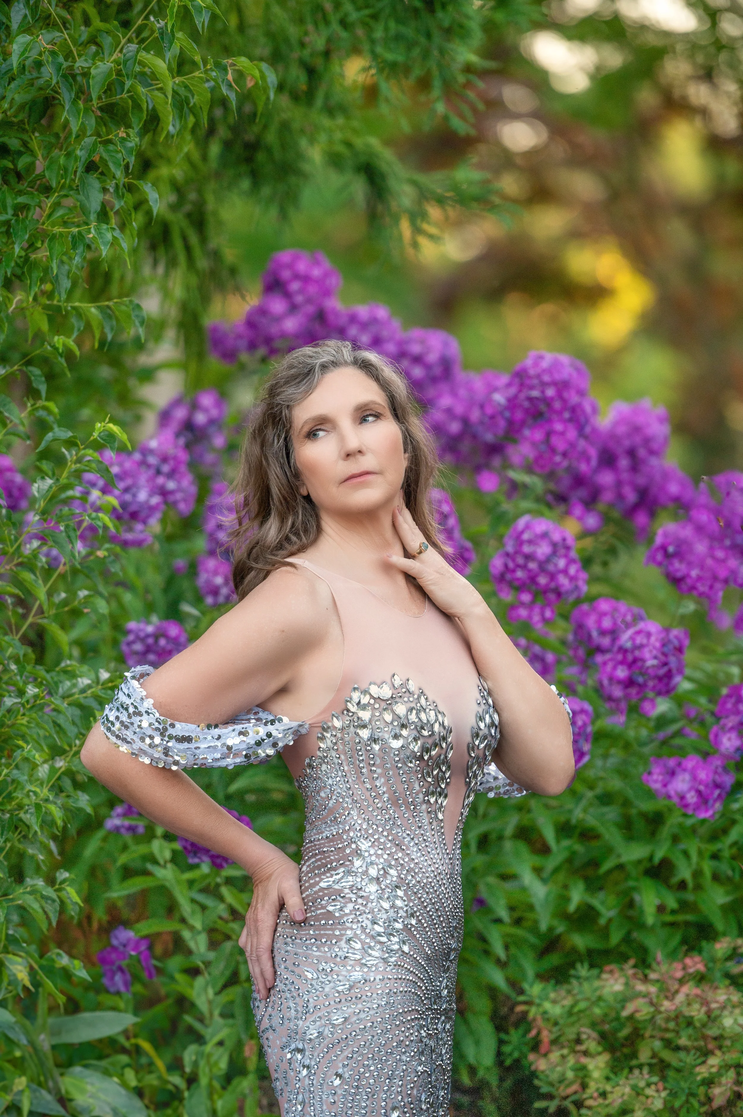 A woman in a sparkly, silver, embellished dress standing outdoors among purple flowers and green foliage, gazing thoughtfully into the distance.