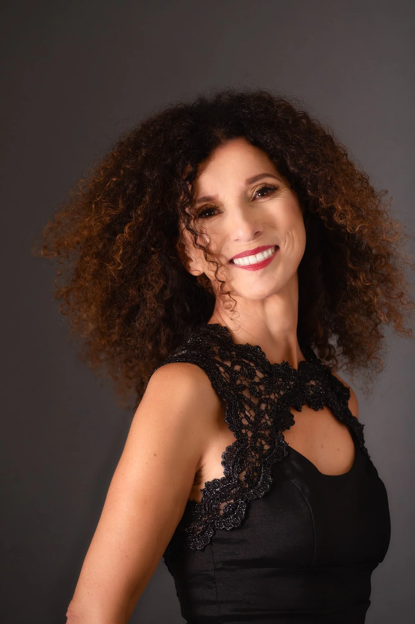 A woman with curly brown hair wearing a black dress with lace detail, smiling against a gray background.