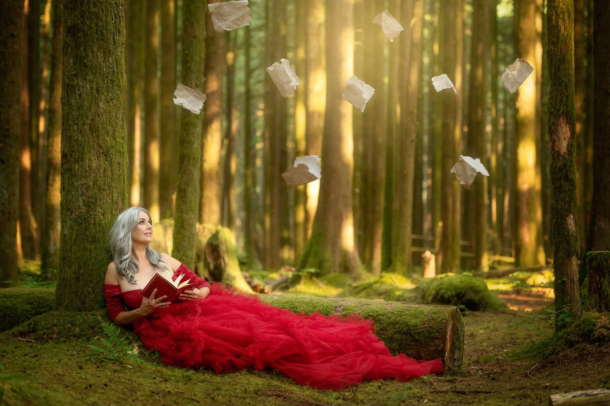 A woman with gray hair wearing a flowing red dress is sitting on a moss-covered log in a dense forest, reading a book, with pages of paper or possibly parchment floating in the air around her, illuminated by sunlight filtering through the trees.