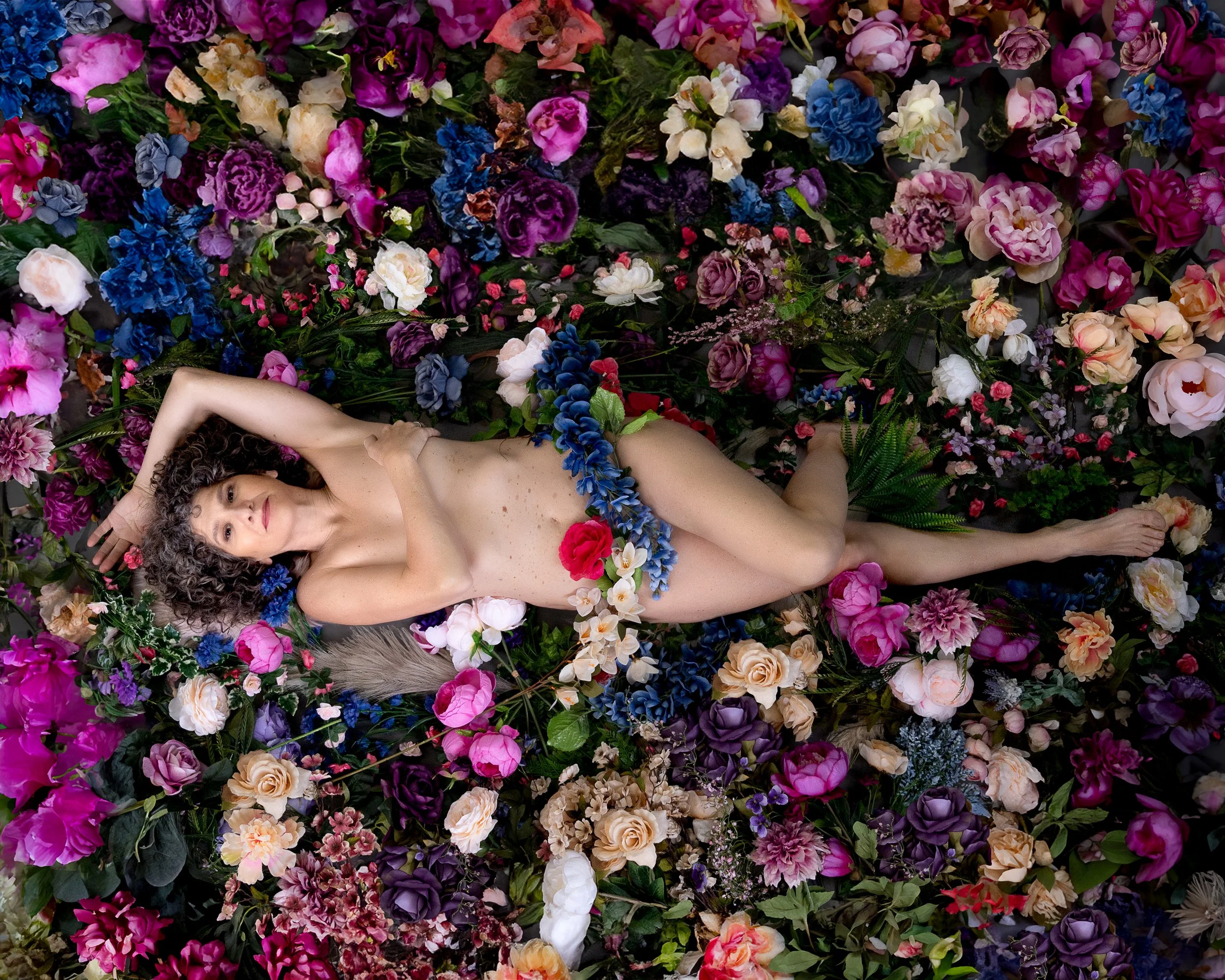 A woman lying amidst an arrangement of colorful flowers, partially covered with flowers, with curly hair, looking at the camera.