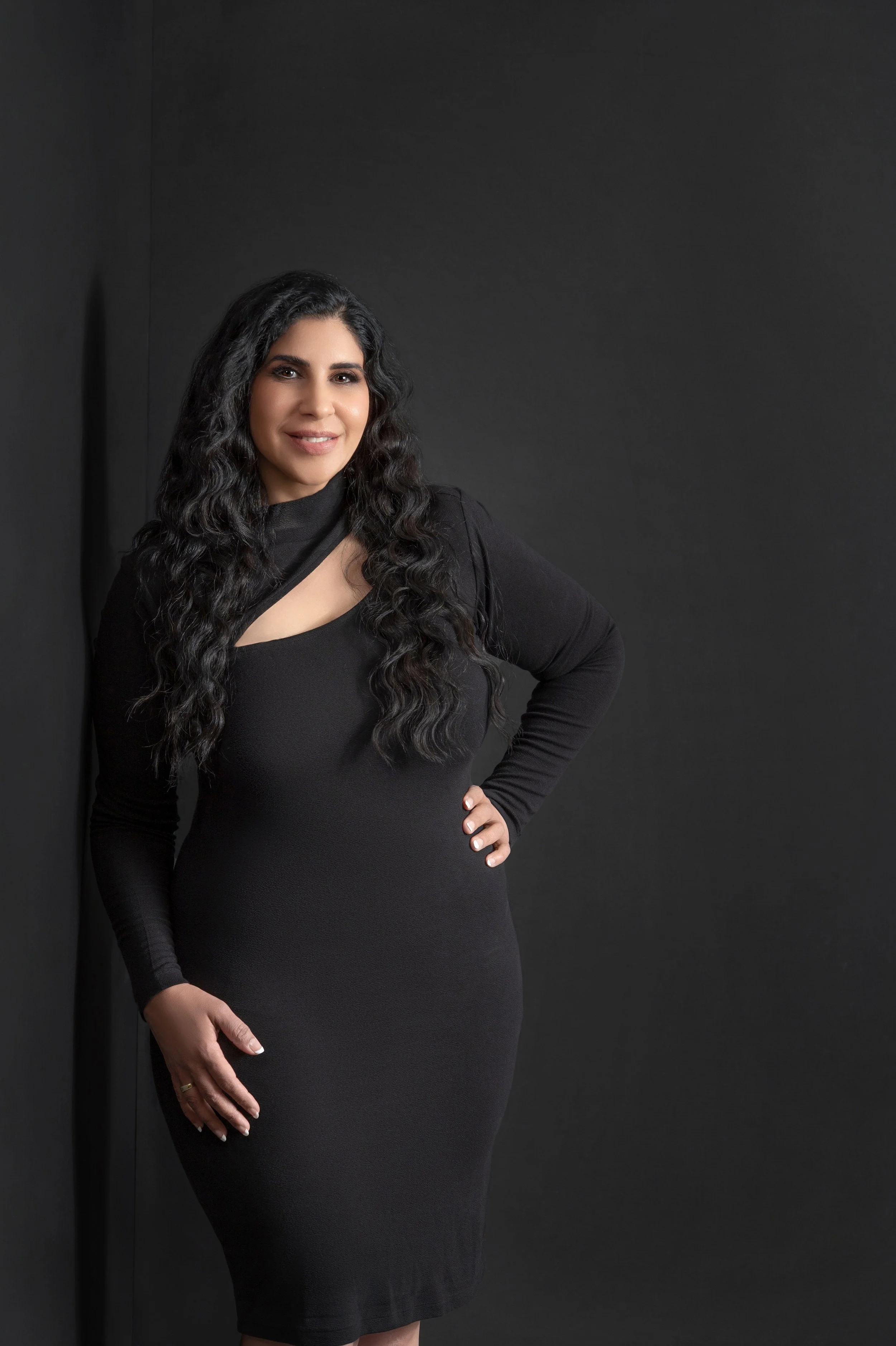 A woman with long, curly dark hair wearing a black dress and standing against a black background.