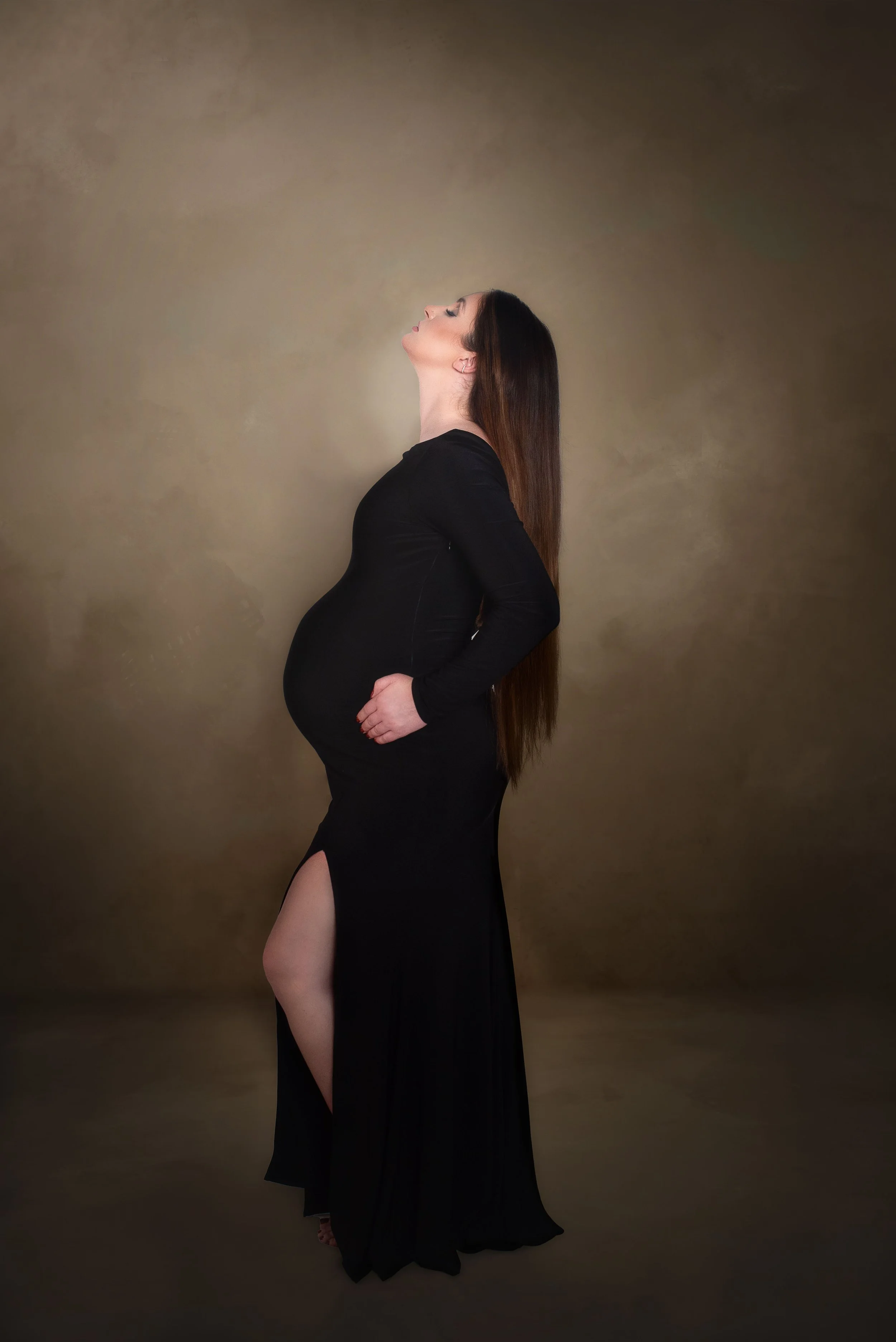 Pregnant woman in a black dress with a slit, standing sideways against a plain brown background, with eyes closed and head tilted back.
