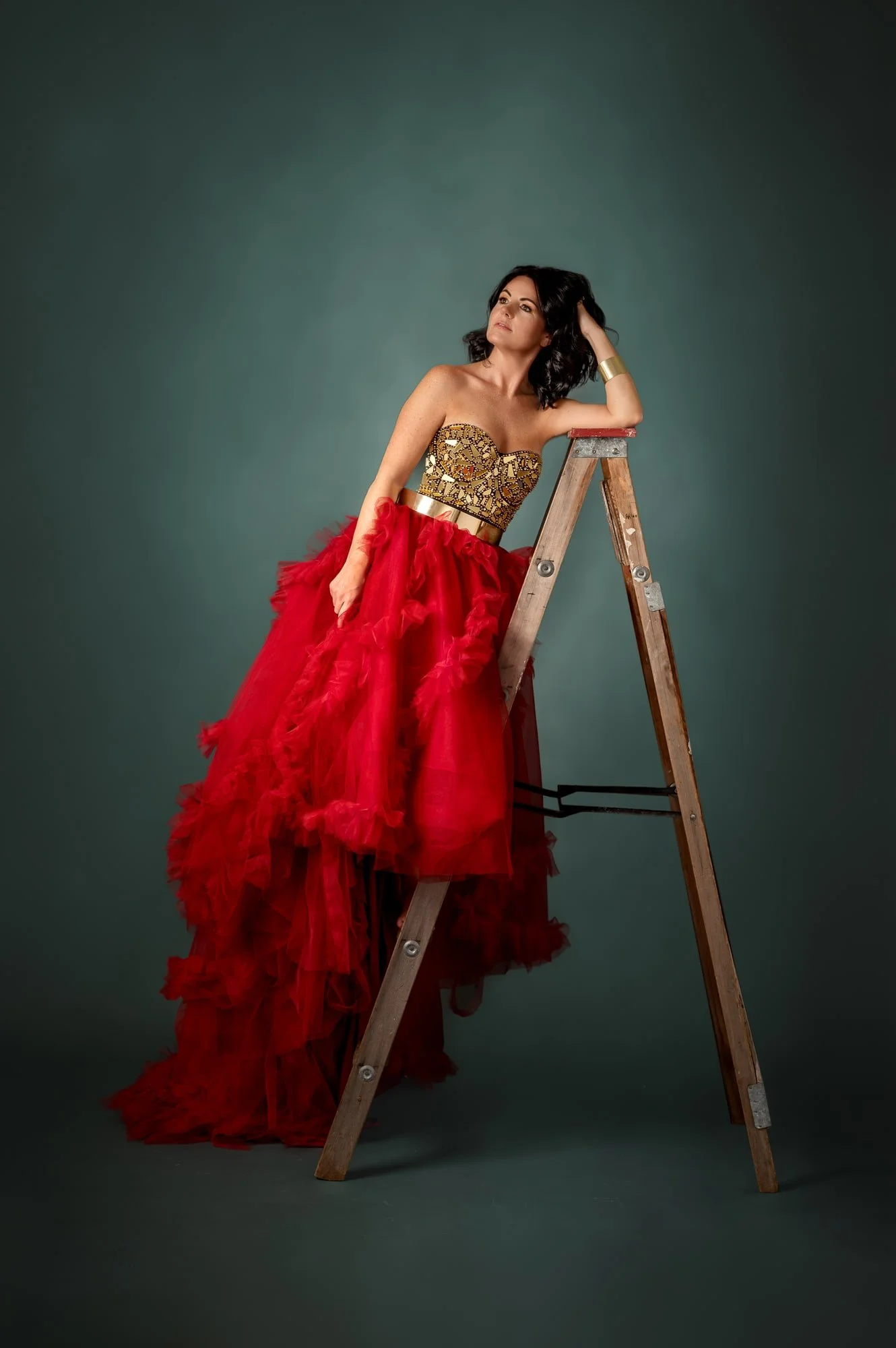 A woman in a glamorous strapless dress with intricate gold and black detailing on the top and a voluminous red tulle skirt, pointing downward, resting her arm on a ladder against a plain background.