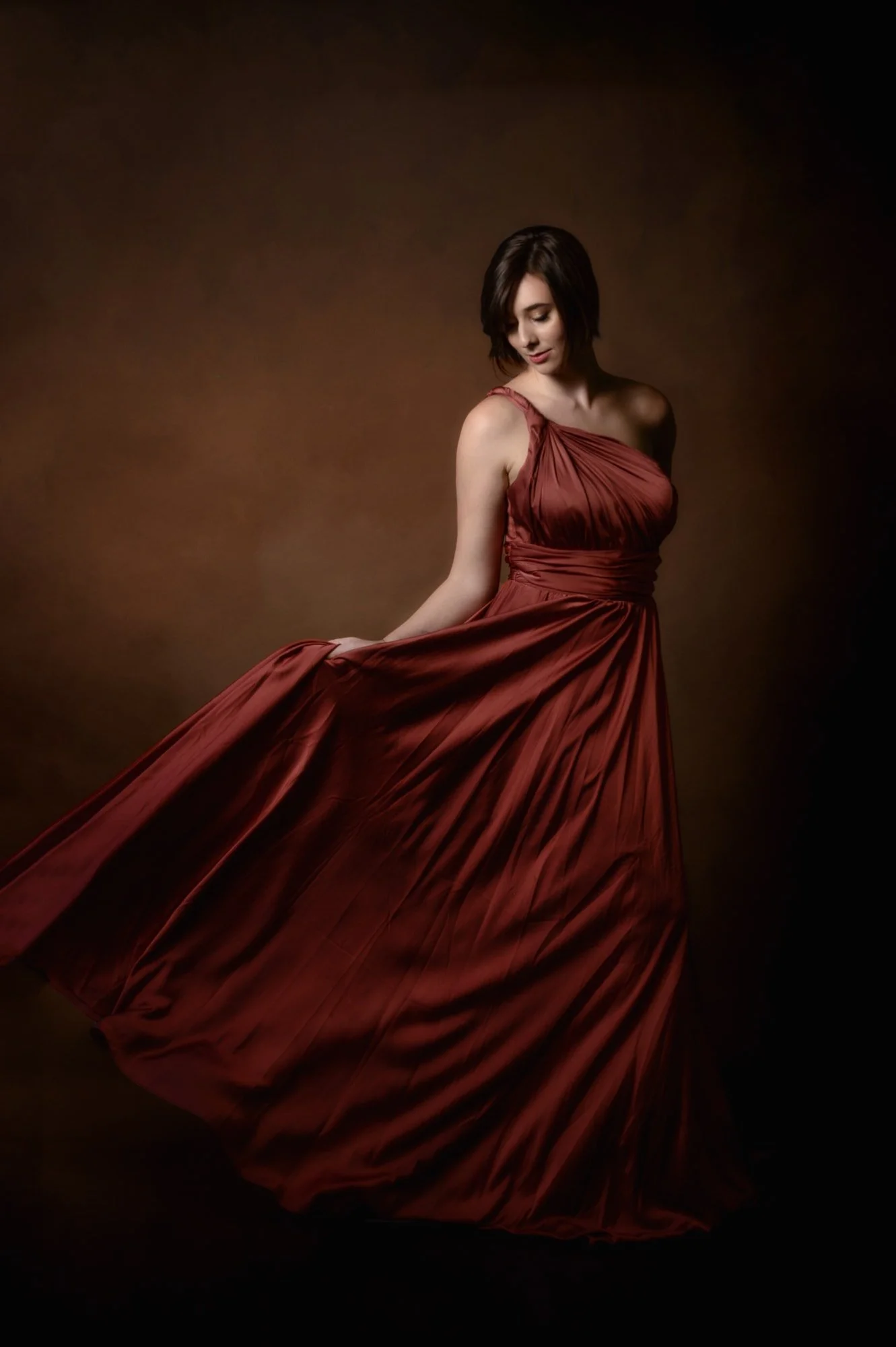A woman in a flowing coral-colored evening gown stands against a brown background, holding the hem of her dress.