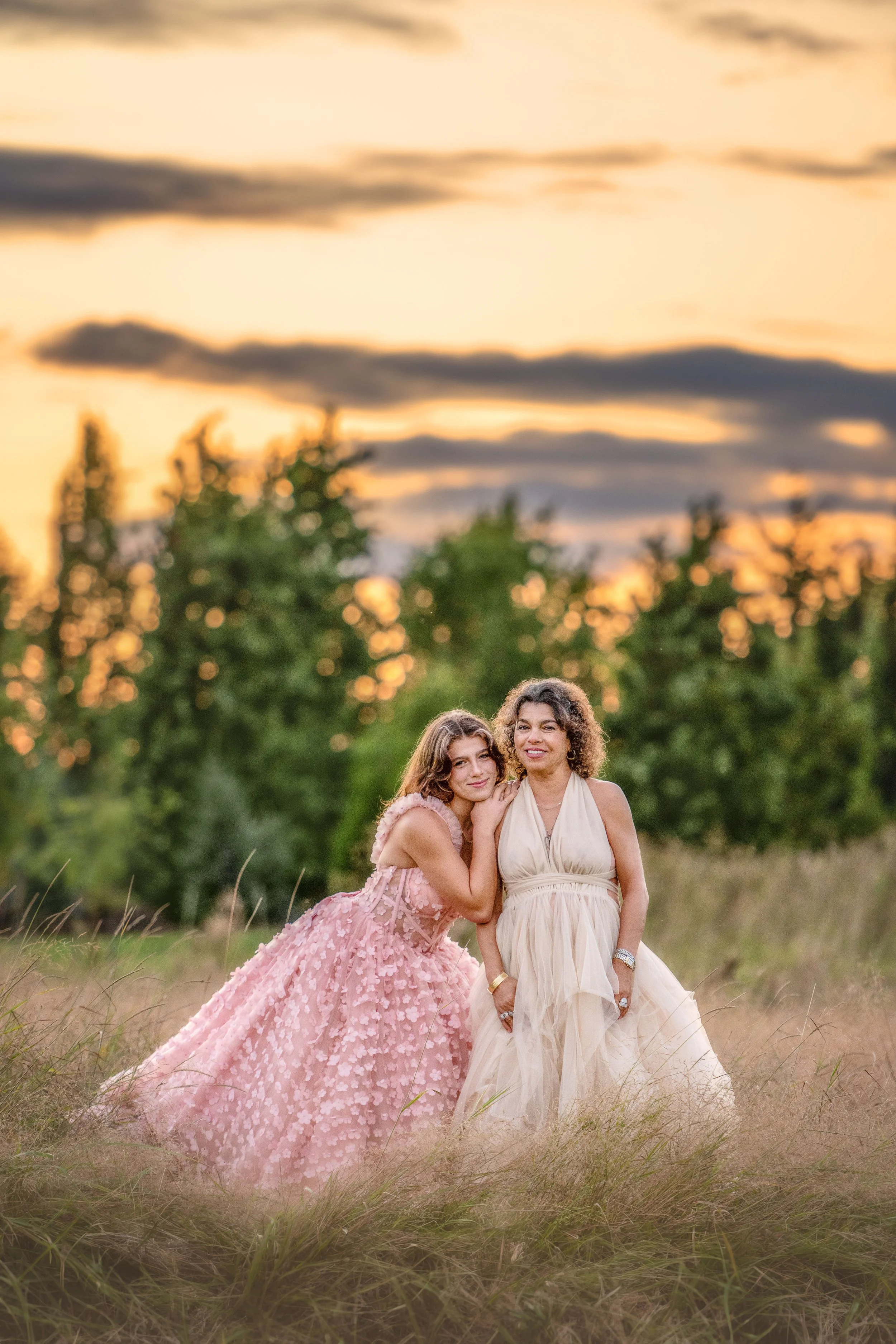Two women in elegant dresses standing in a grassy field during sunset, with trees in the background.