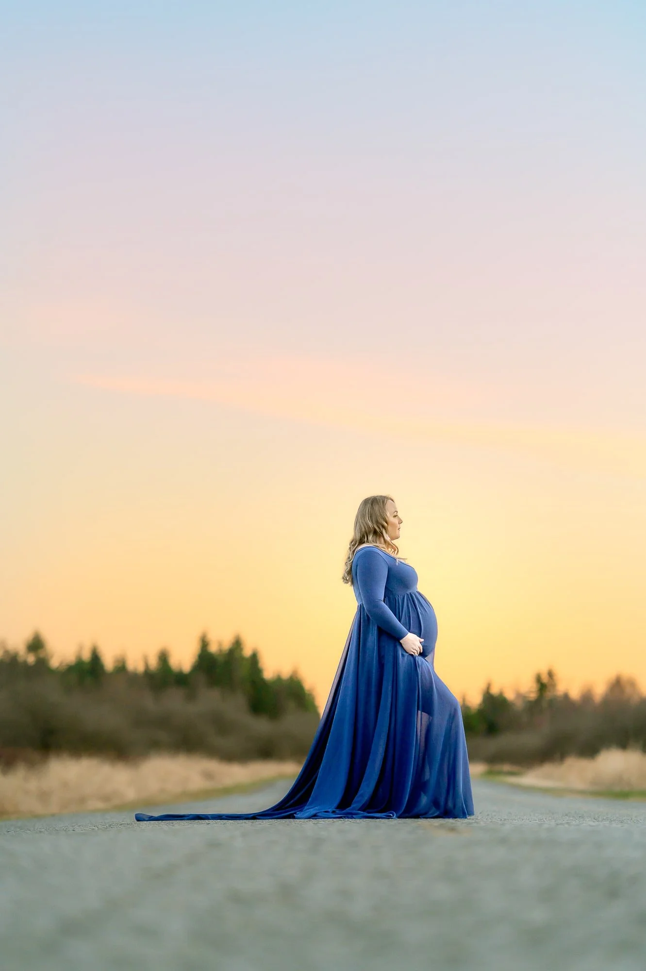 Pregnant woman in a flowing blue gown standing on an empty road at sunset.