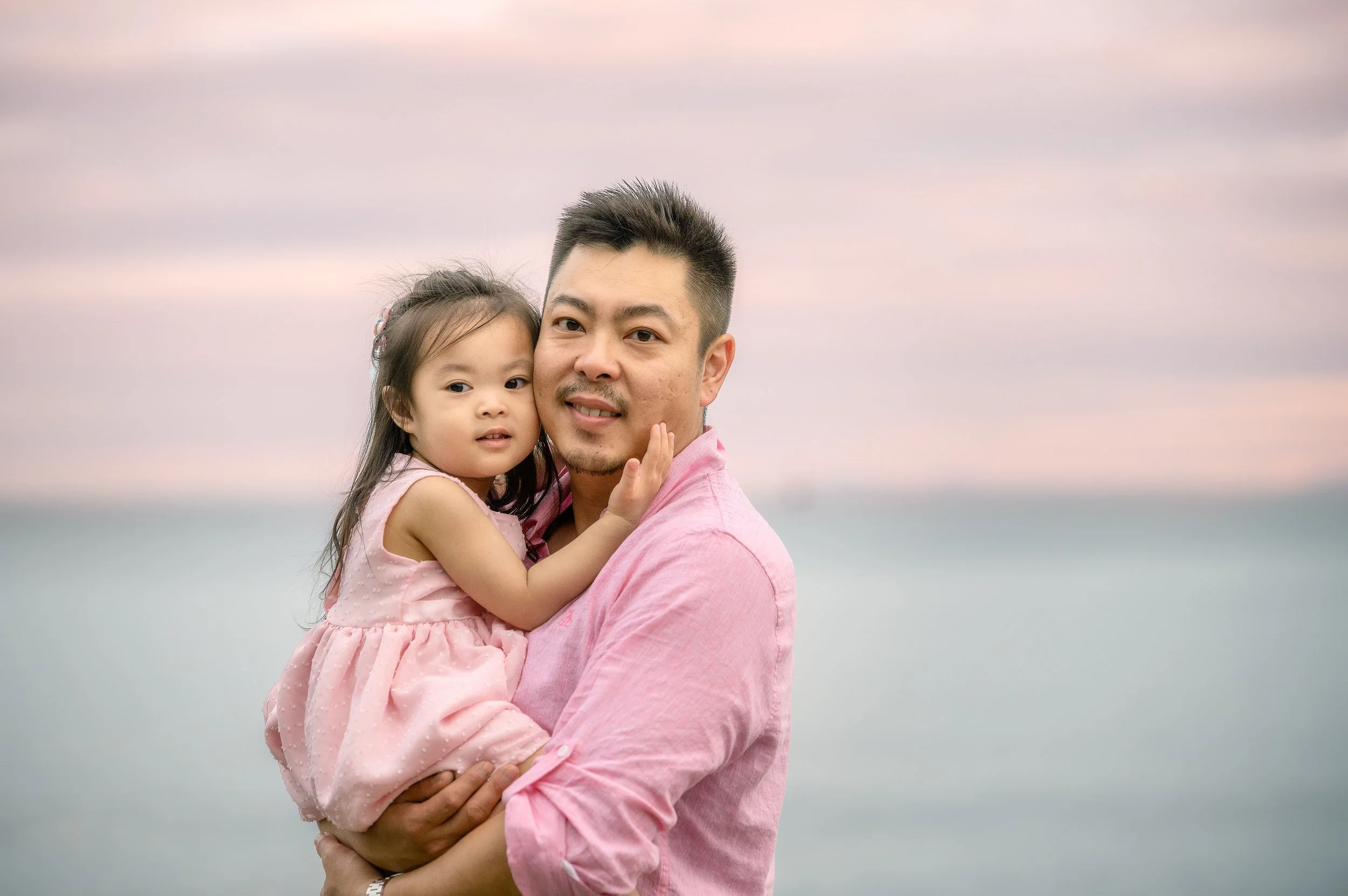 A man and a young girl at the beach during sunset, with the man holding the girl in his arms. Both are wearing pink clothing and looking at the camera.