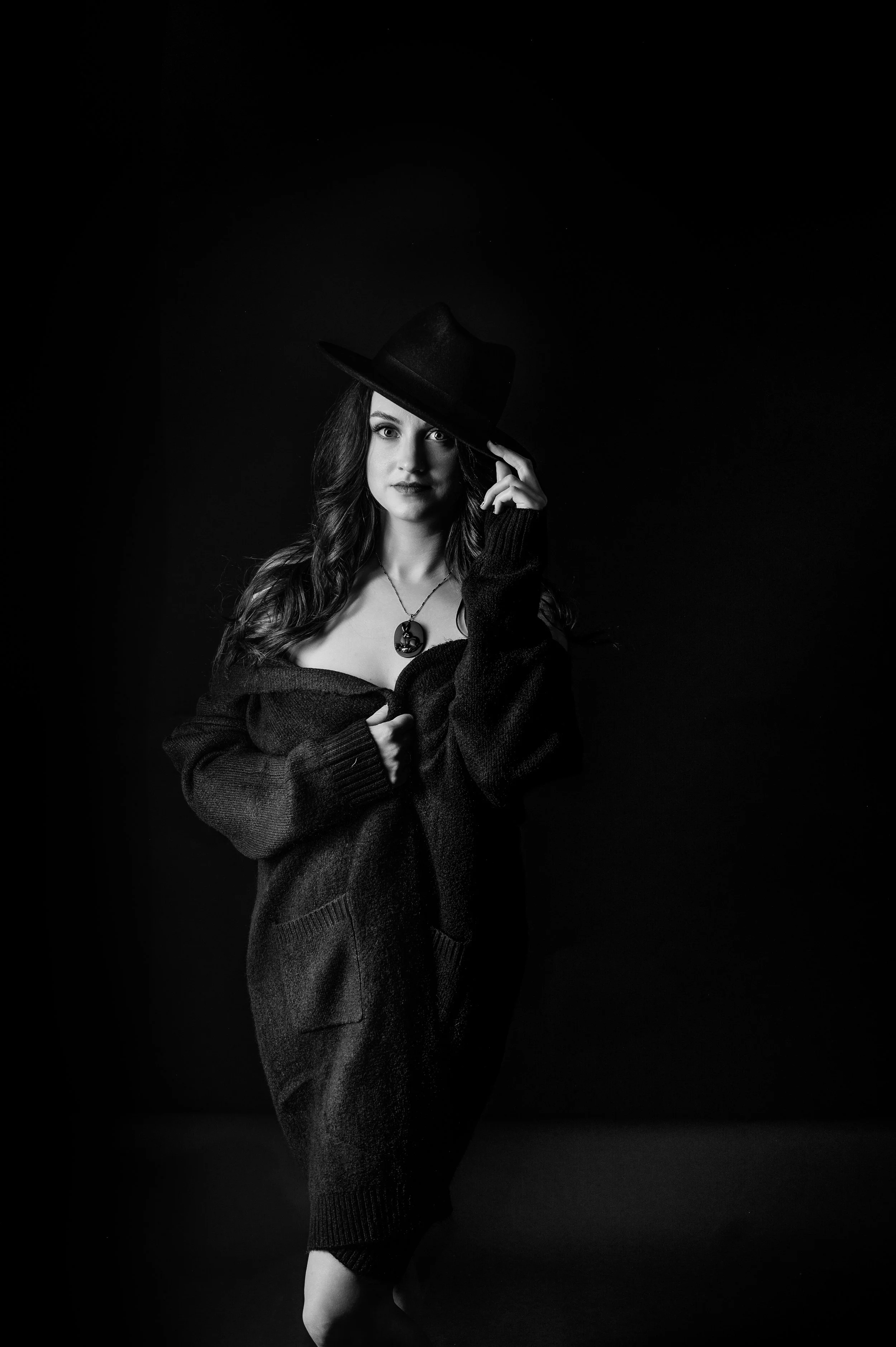 A black and white portrait of a woman wearing a wide-brimmed hat, an off-the-shoulder sweater, and a necklace, standing against a dark background.