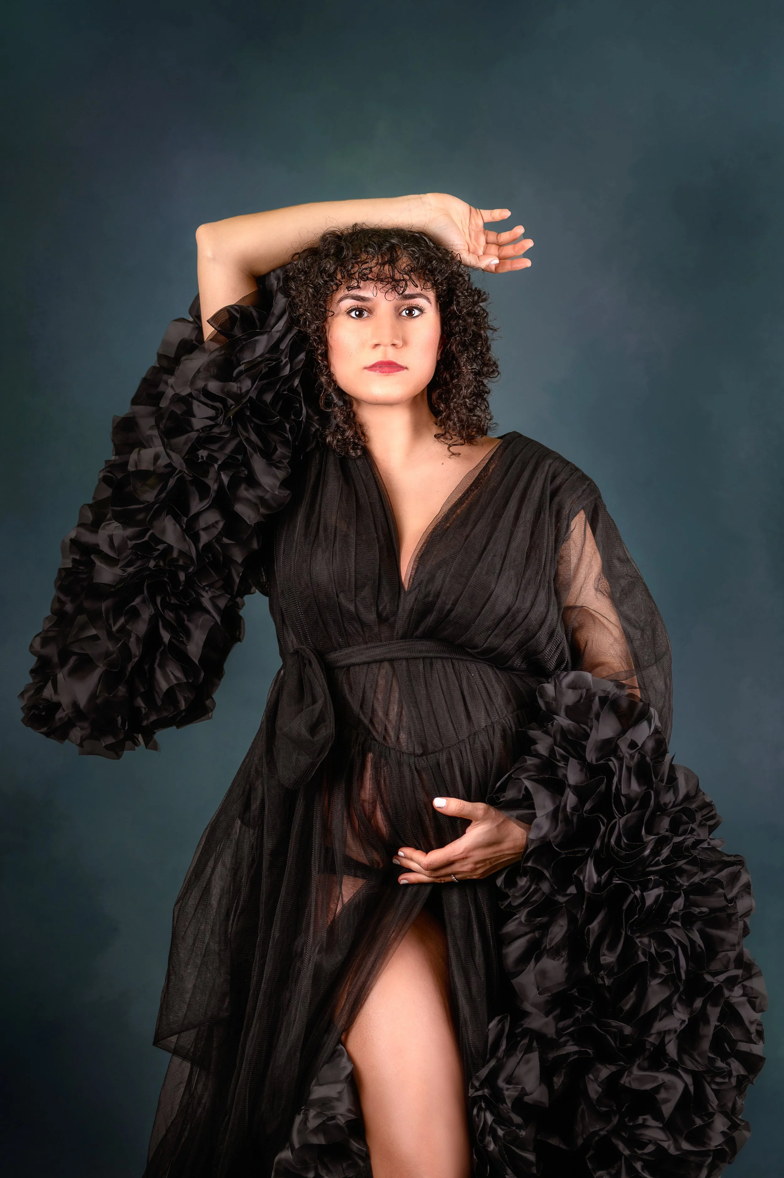 A pregnant woman with curly dark hair posing with one hand on her belly and another raised above her head, wearing a black sheer dress with ruffled sleeves against a dark green background.