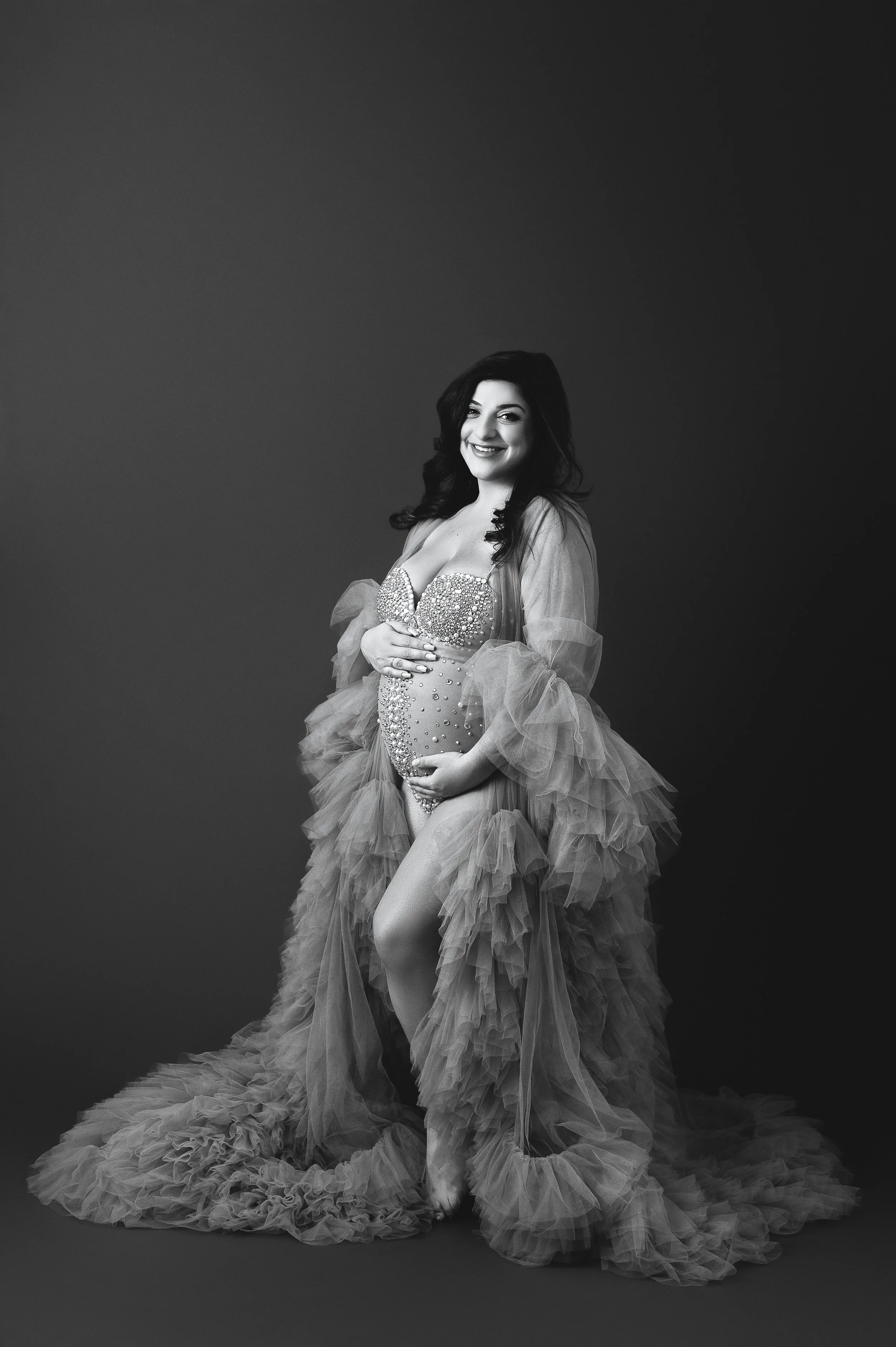 A pregnant woman with long, dark wavy hair, wearing a glamorous, bejeweled dress with puffy, layered tulle fabric, standing against a plain dark background.